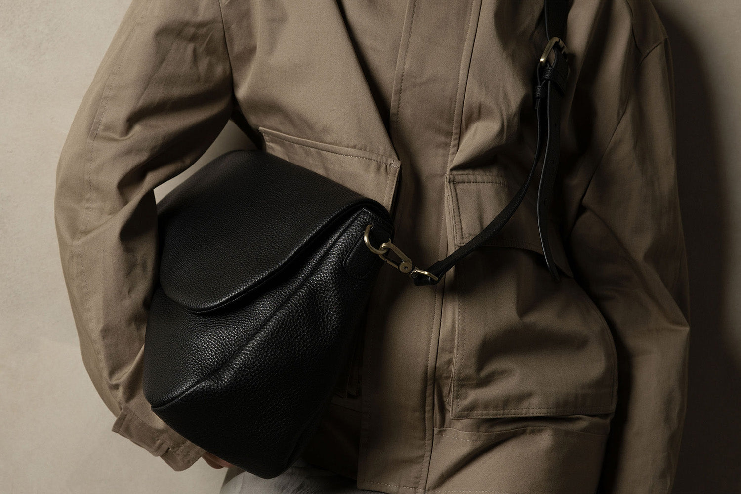 A person wearing a light brown jacket carries a black leather crossbody bag with a flap closure and gold-tone hardware, shown against a neutral background.