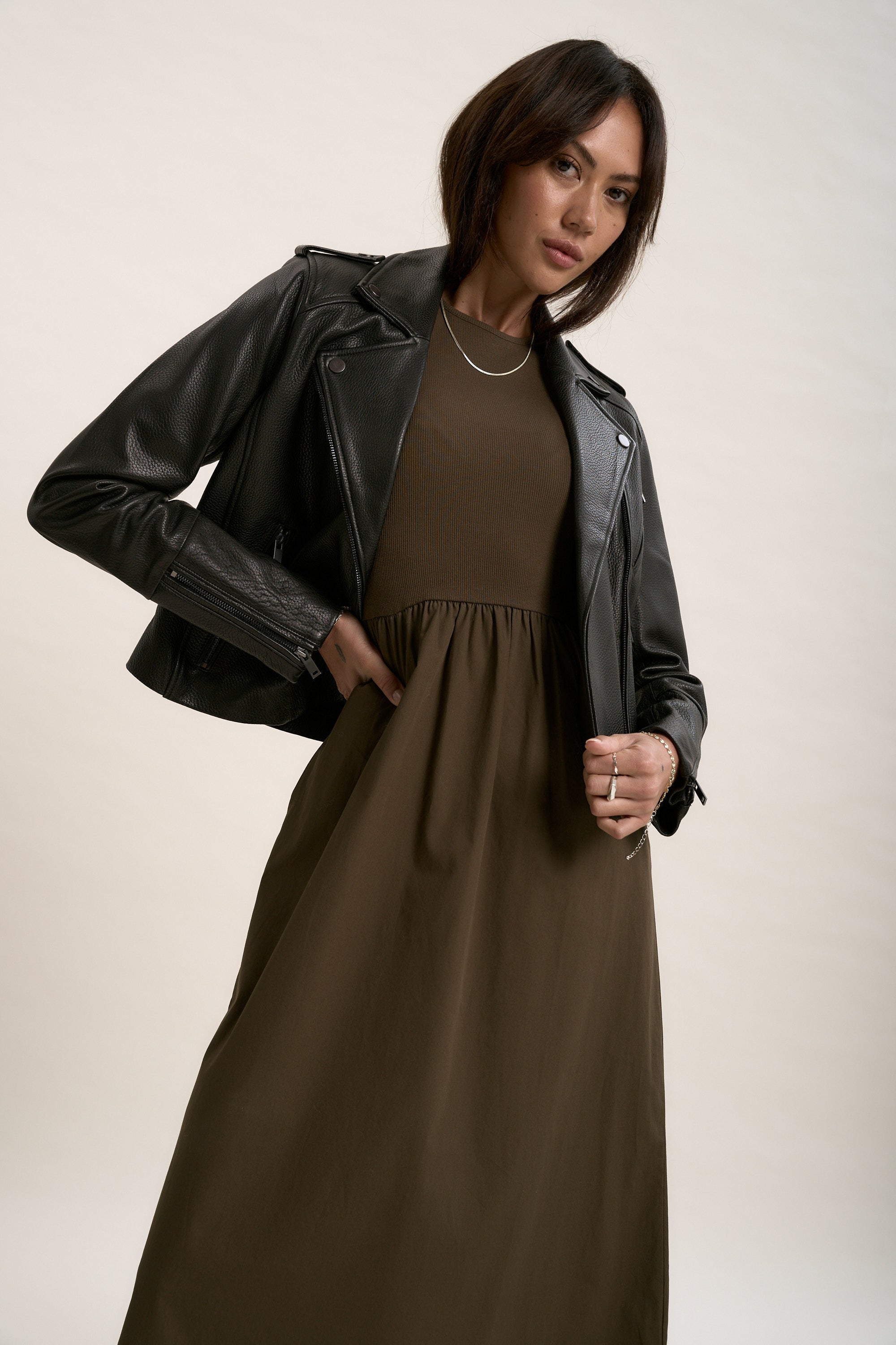 A woman poses confidently with one hand on her hip, wearing Max's The Perfect Leather Biker Jacket over a brown dress—a timeless staple for any woman's wardrobe—set against a plain light background.