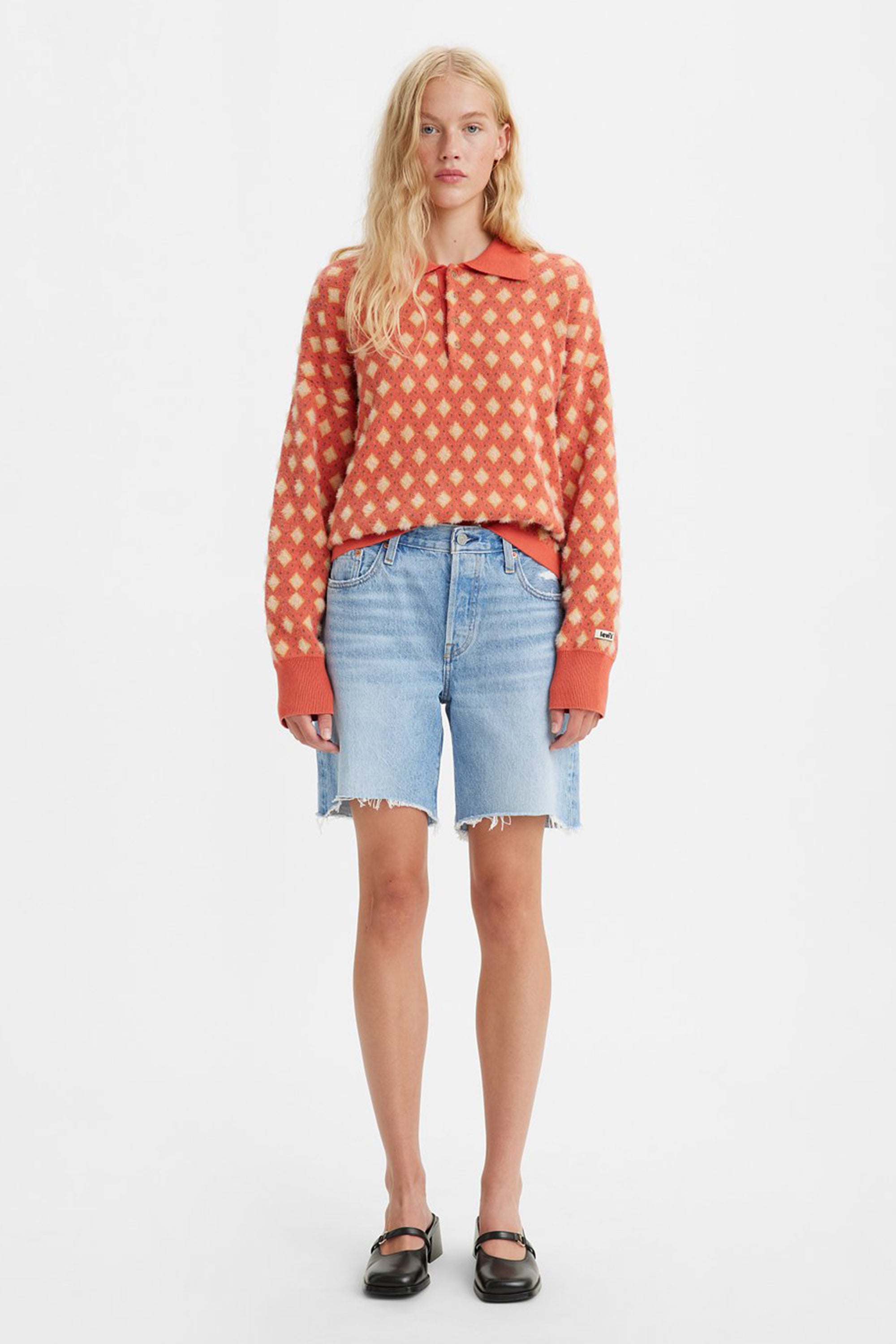 A woman faces forward in a long-sleeve orange and cream checkered sweater, Levi's 90's 501 Short vintage denim shorts with frayed hems, and black Mary Jane shoes, set against a plain white background.