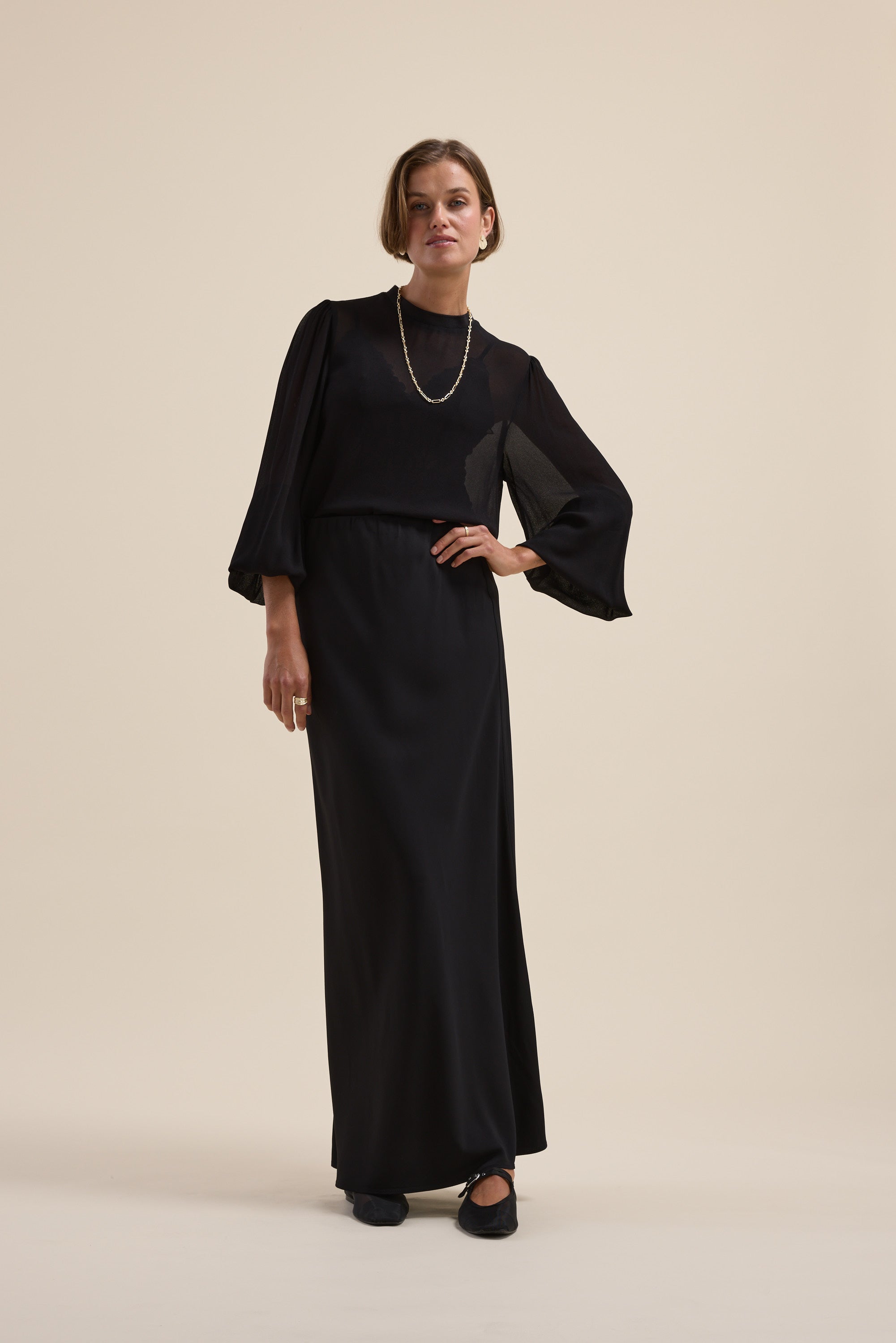 Woman wears Max’s The Ultimate Layering Top in sheer black, posing against a beige background.