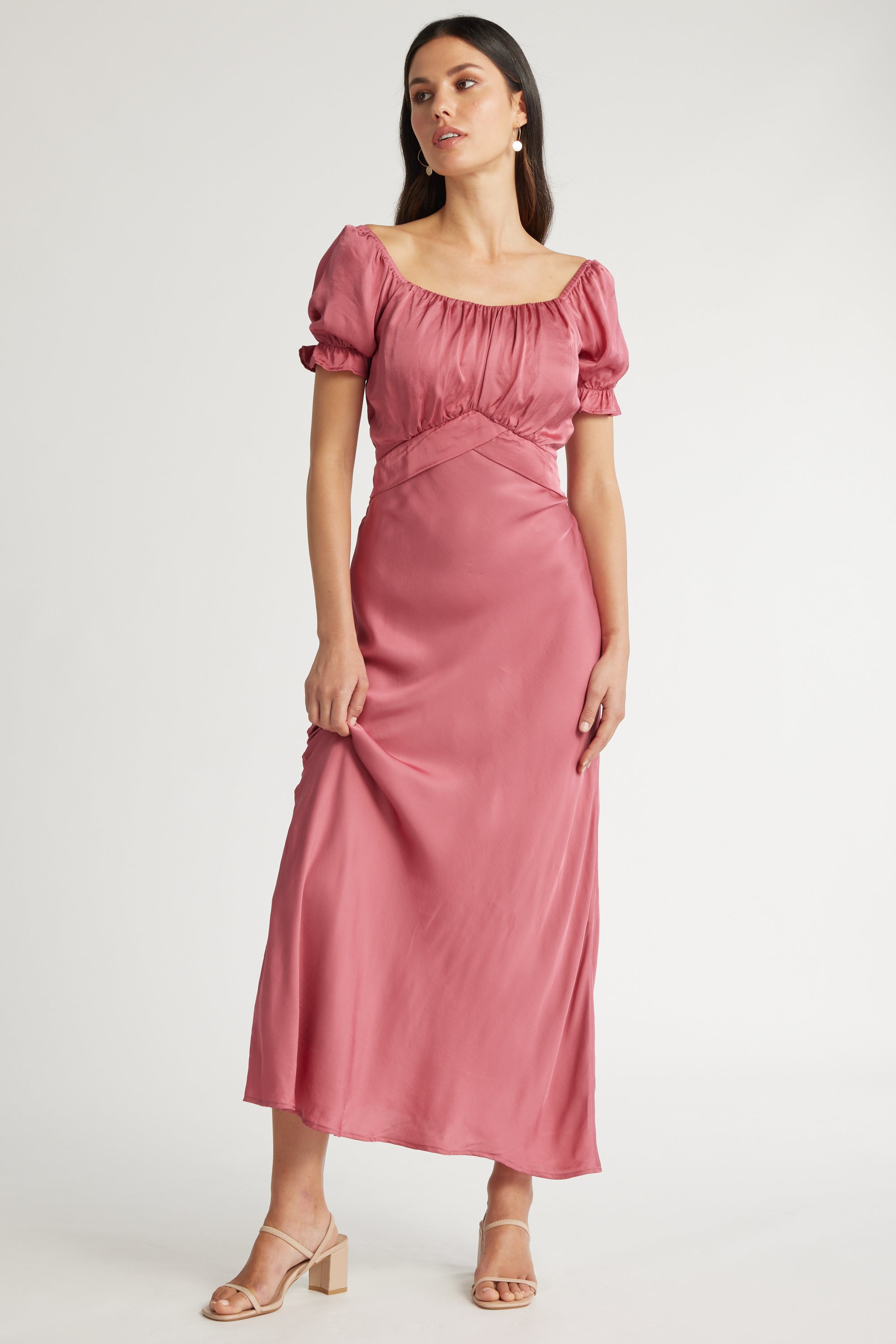 A woman in the Sara Puff Sleeve Dress by Max, featuring a pink maxi length and square neckline, paired with nude block-heeled sandals, stands against a plain white background.