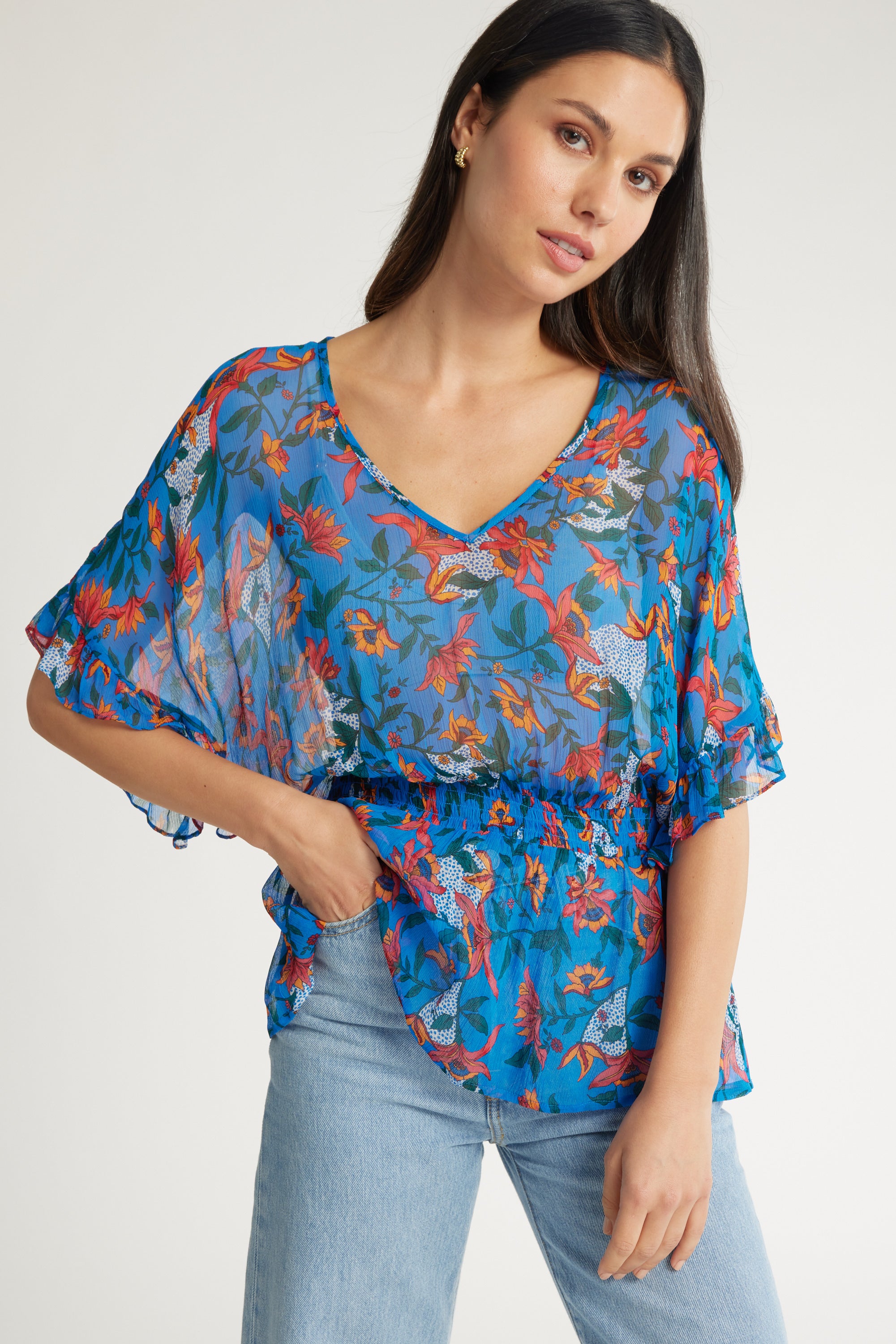 A woman wearing the Max Selena Frill Detail Top—a blue sheer floral print with frills and short flowy sleeves—paired with light blue jeans, stands with one hand in her pocket against a plain background.