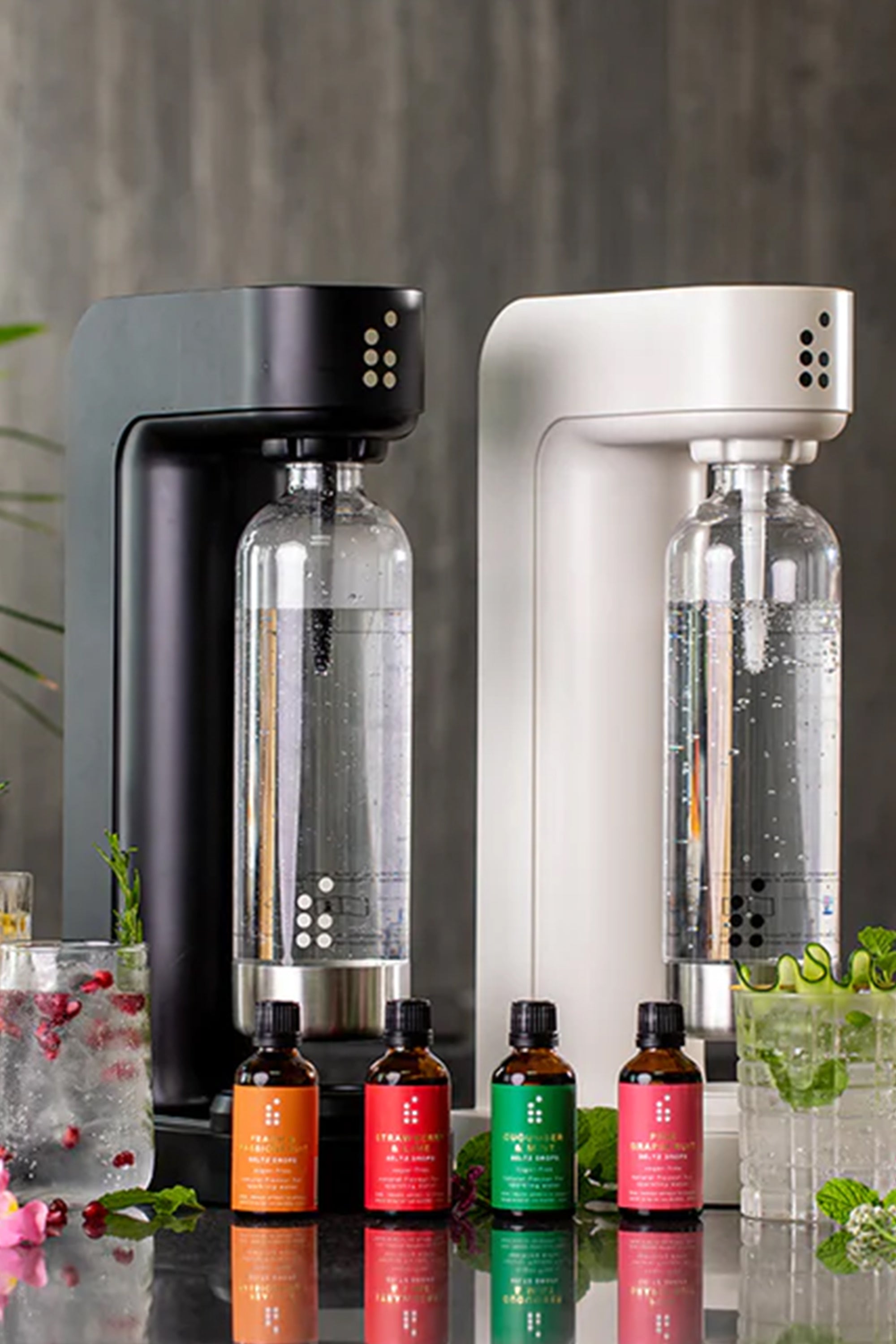 Two sleek sparkling water makers stand side by side with bottles of sparkling water. In front are four Six Barrel Soda Company Seltz Drops and glasses garnished with fruit and mint for low-waste, sugar-free drinks.