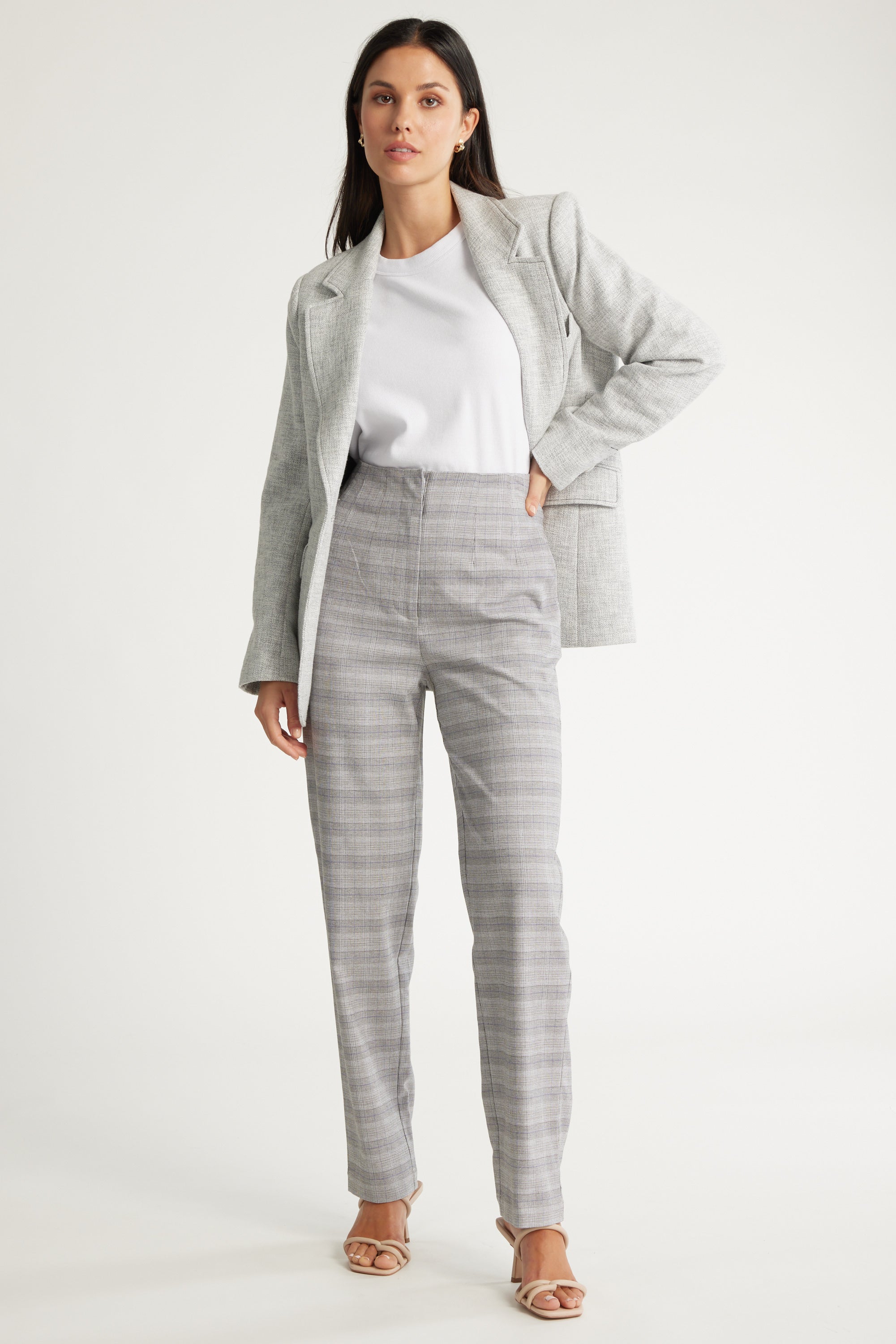 A woman poses with one hand on her hip, wearing Max's Nyla Tapered Leg Pant in light gray check with a matching blazer, white t-shirt, and beige heeled sandals against a plain background.