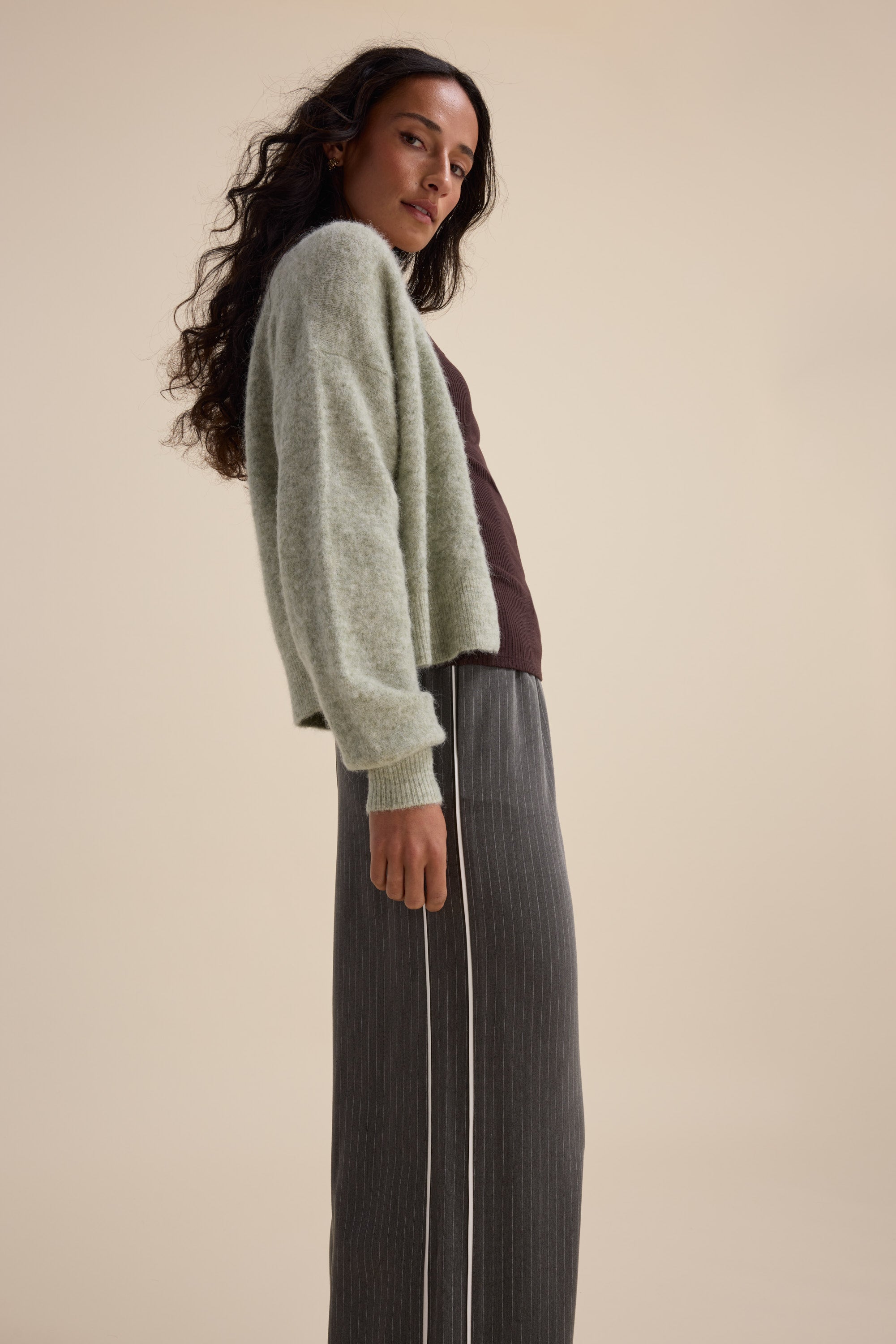 Woman in The Ultimate Luxe Cardigan by Max, dark top, striped gray pants, standing sideways.