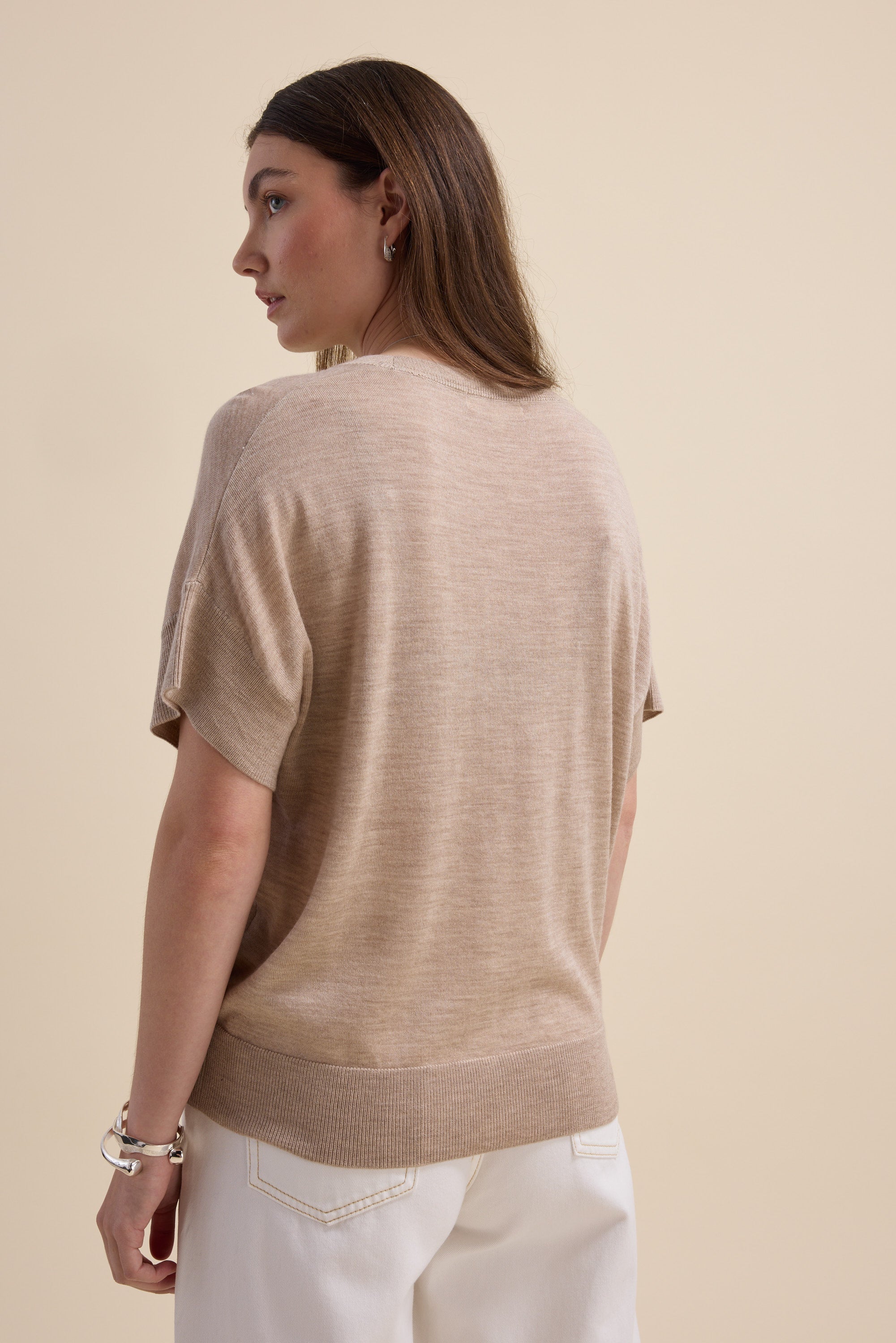 Woman wears Max's The Merino Silk Knit Tee with white pants, facing away on beige background.