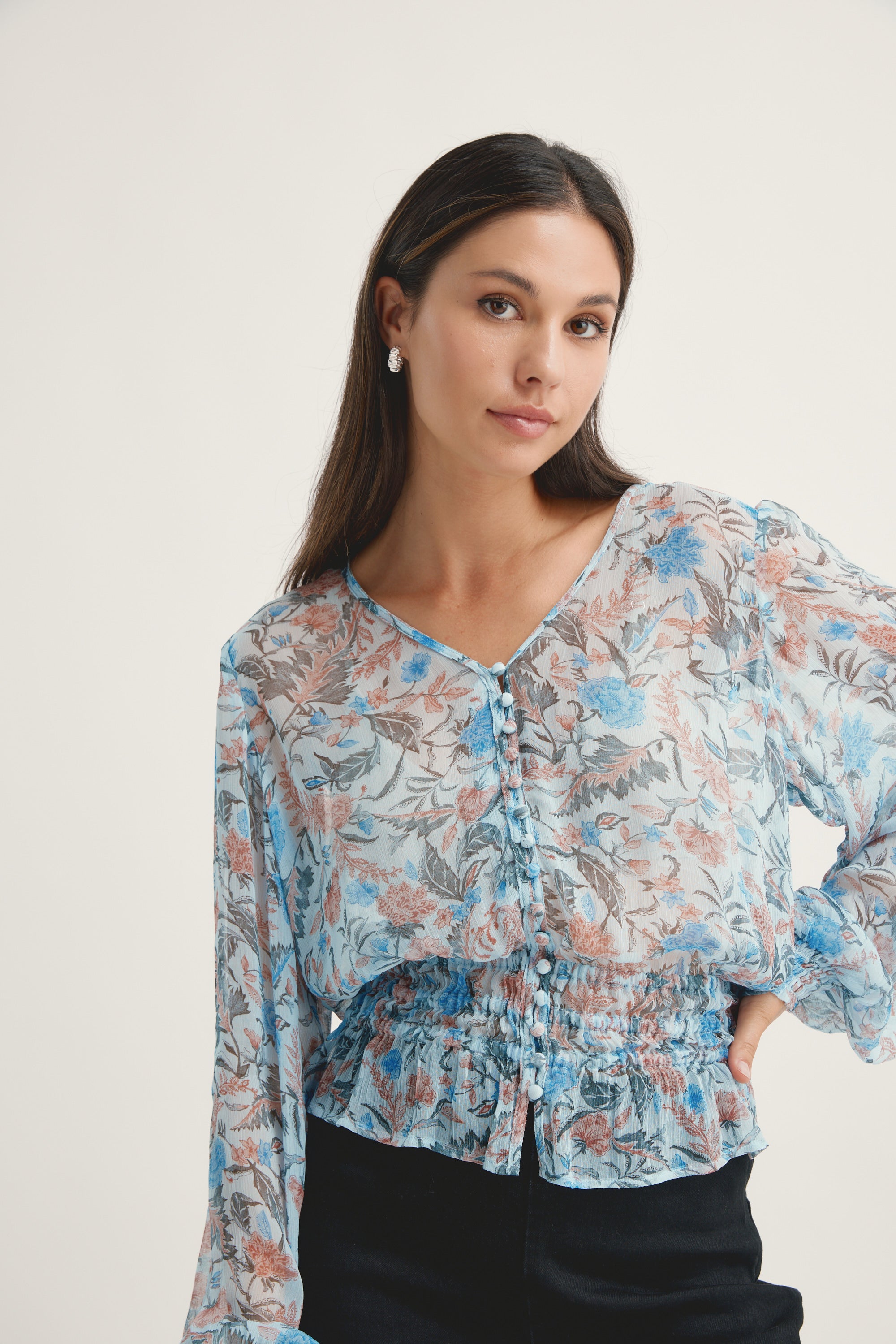 A woman with long brown hair models the Max Joan Button Front Blouse, a sheer floral print in blue and beige with ruffle details, standing against a plain light background and gazing slightly to the side.
