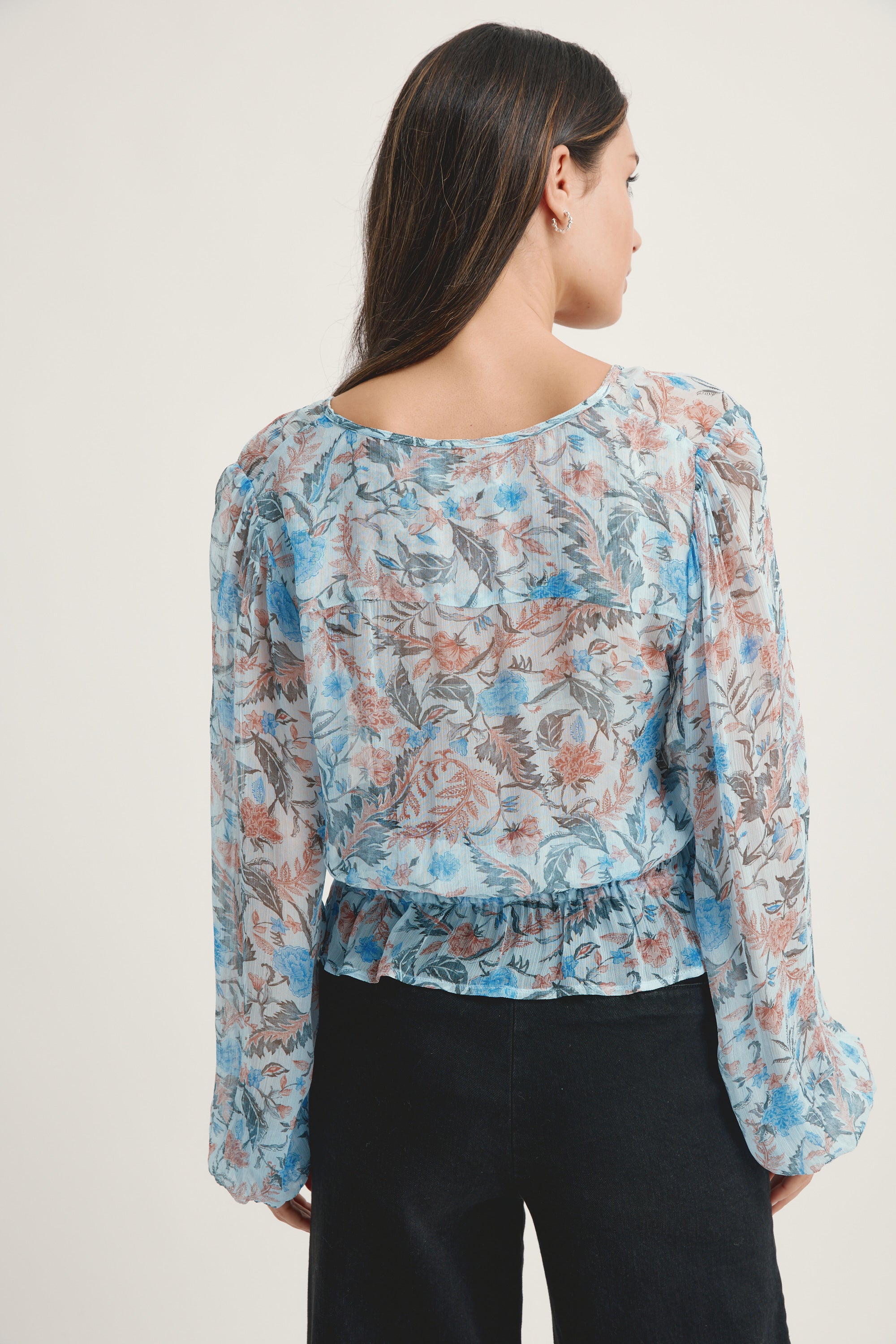 A woman with long dark hair, seen from behind, wears the Max Joan Button Front Blouse—a sheer, long-sleeve floral top with a loose fit—paired with black pants against a plain light background.