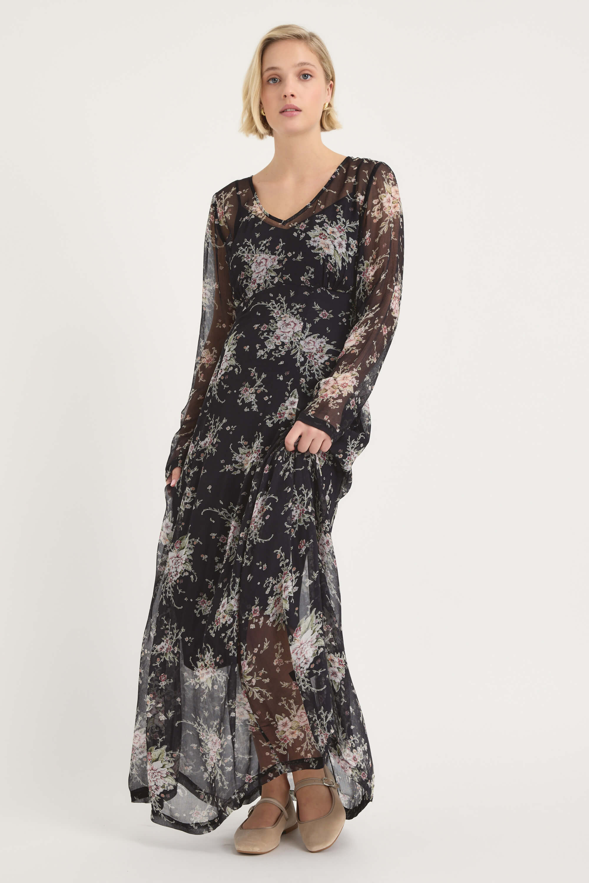 A woman with short blonde hair, wearing tan shoes, models the Amber Long Sleeve Dress by Max— a sheer floral maxi dress with long sleeves and a V neckline—against a white background, looking slightly to the side.