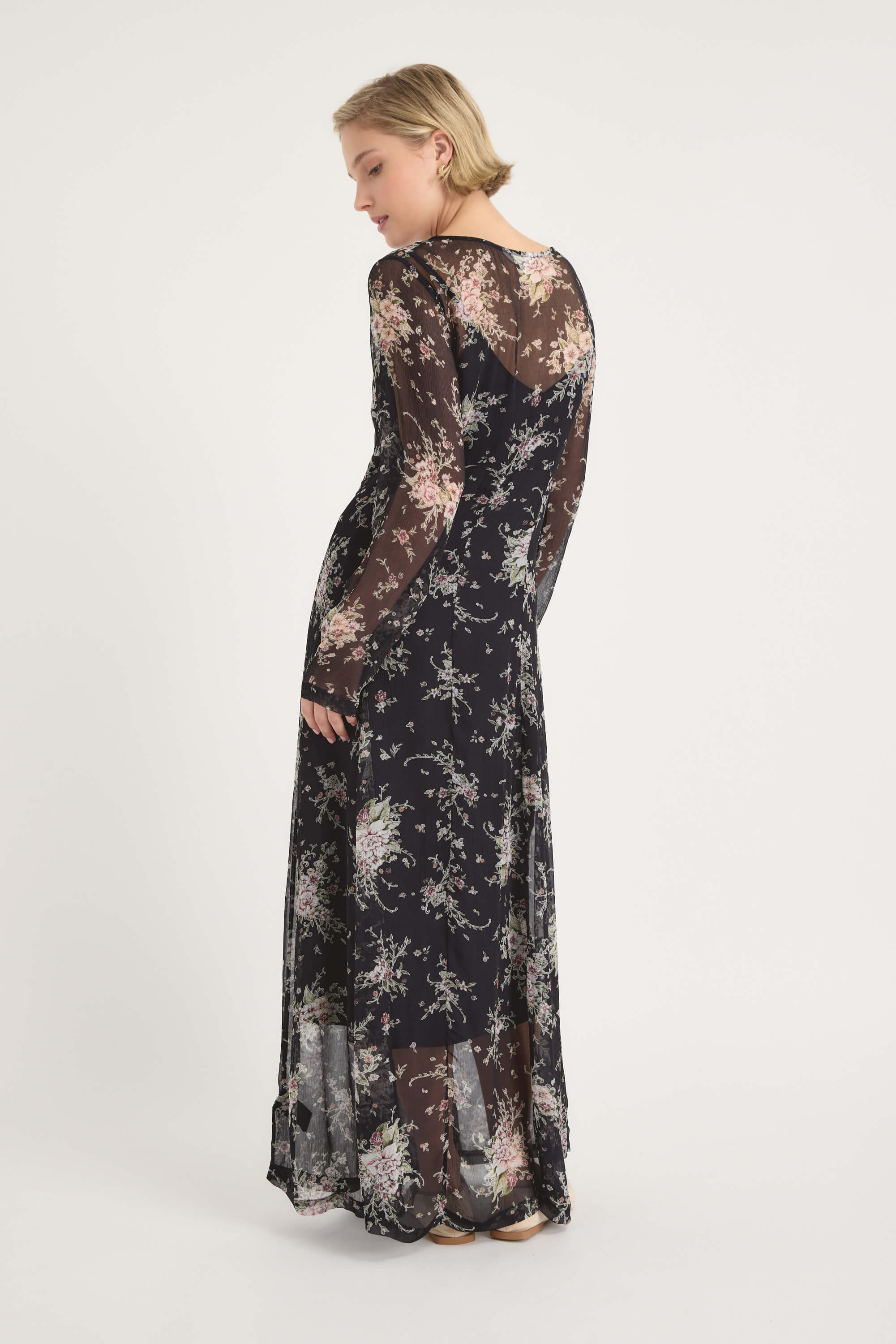 A woman with short blond hair stands sideways in the Amber Long Sleeve Dress by Max, a black sheer floral maxi with a flowy skirt and long sleeves. She is set against a plain white background.