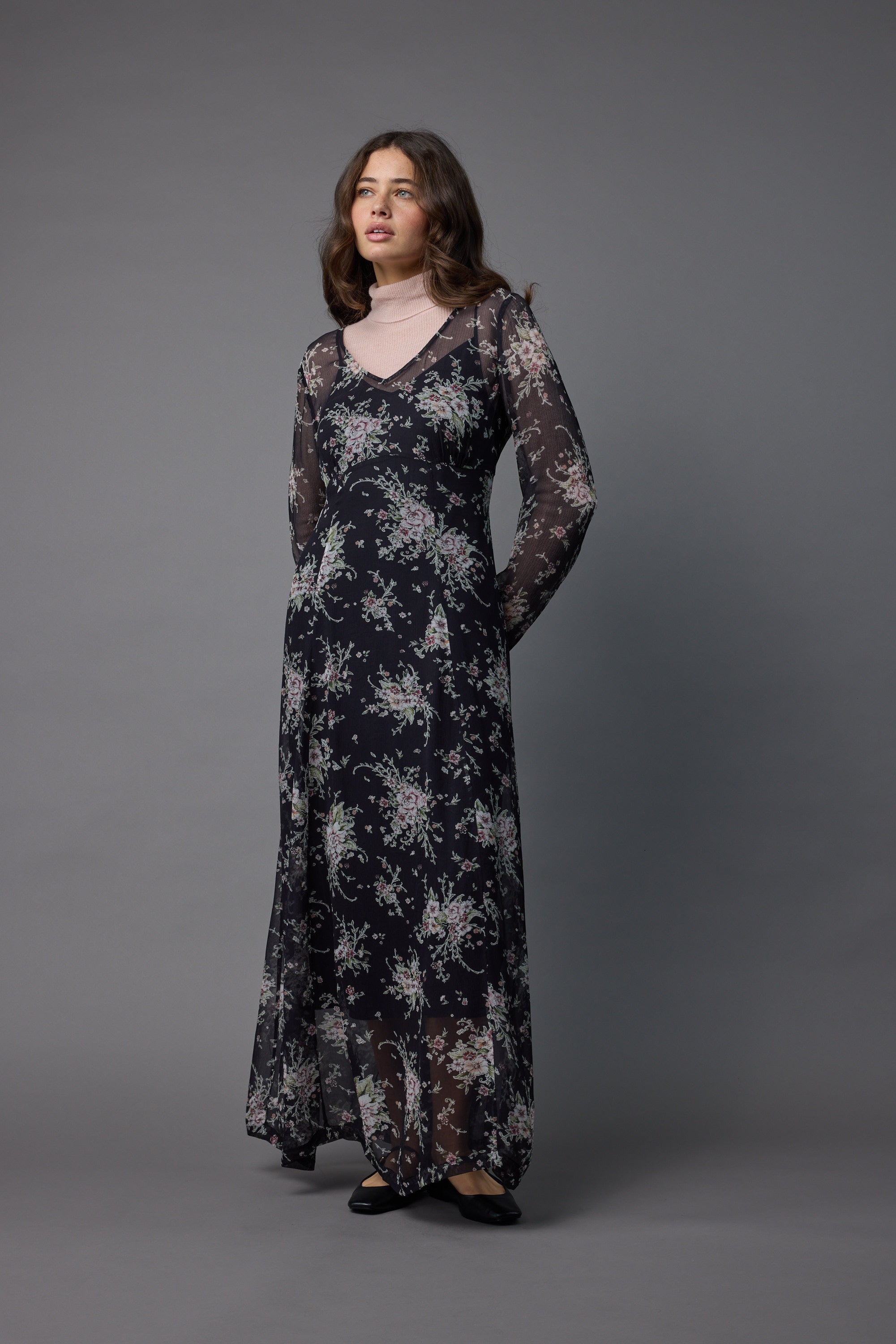 A woman with long wavy brown hair stands against a gray background, wearing the Max Amber Long Sleeve Dress over a pale pink turtleneck, gazing slightly to the side.