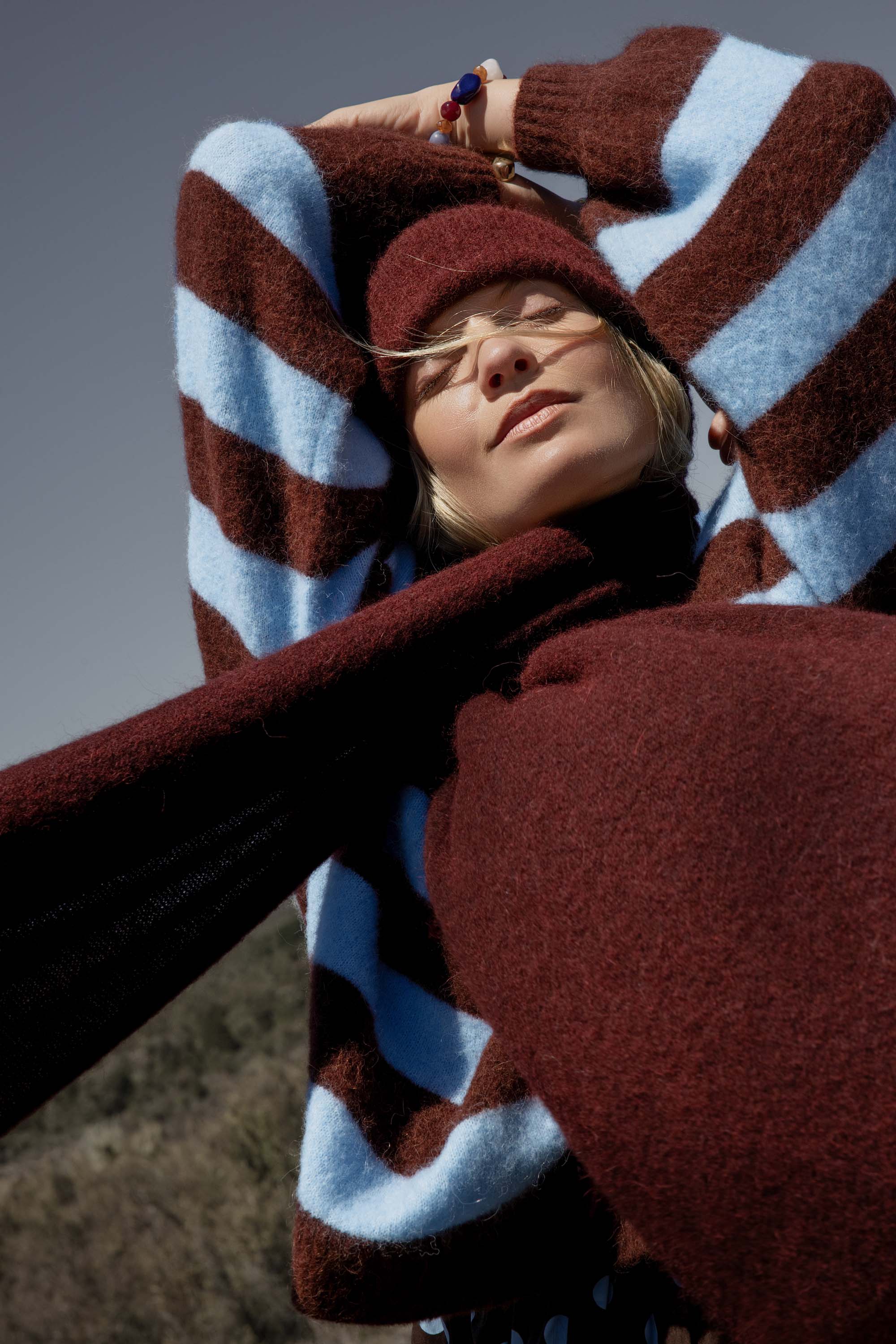 Woman poses outdoors in Max’s Hailey Luxe Whisper Knit Scarf, arms raised and eyes closed in sunlight.