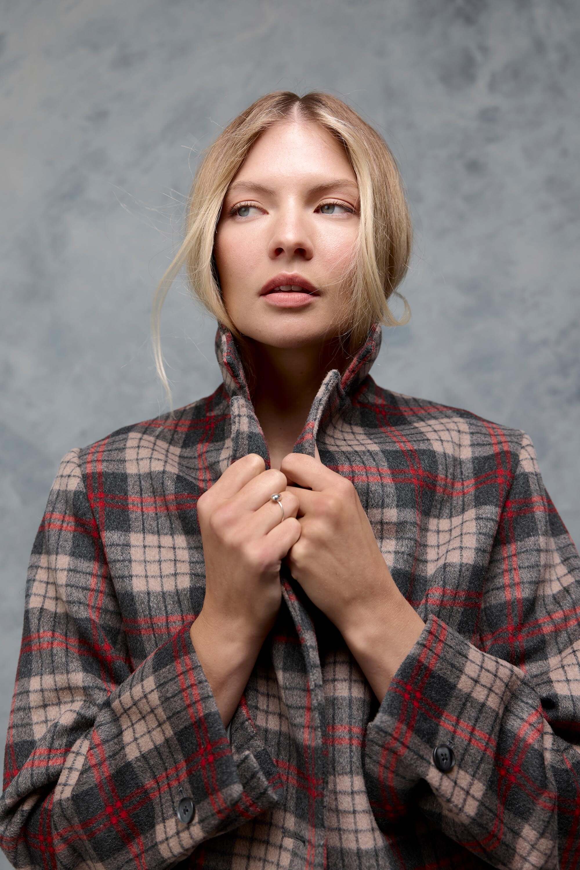A blonde woman poses against a gray textured background, wearing the Max Mackenzie Coat—an Inverness check wool blend with red, black, and beige patterns—holding the collar up and looking left; a standout piece for any winter wardrobe.