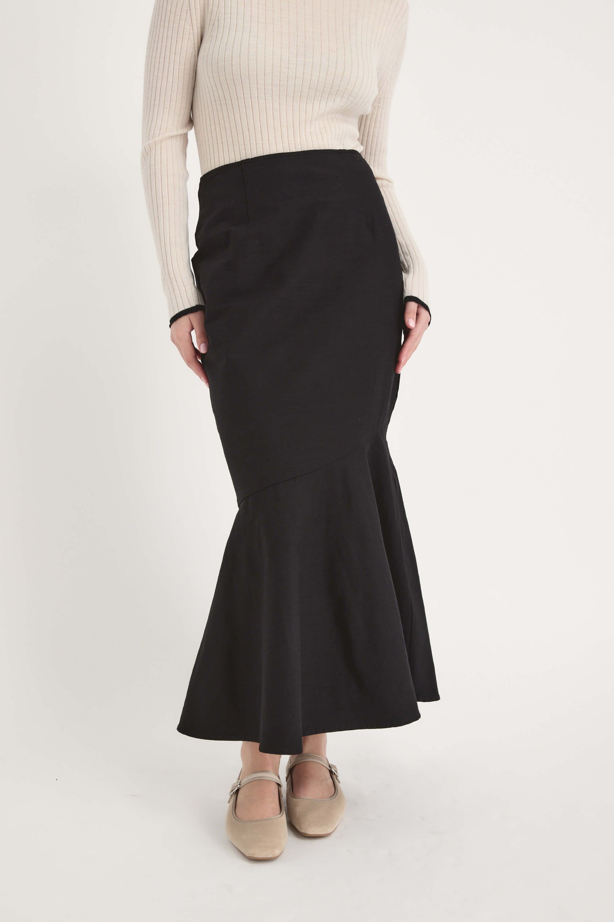 A person wearing a cream ribbed long-sleeve top, the Frances Flared Skirt by Max—a high-waisted black skirt with a fishtail hem—and beige shoes stands against a plain white background.