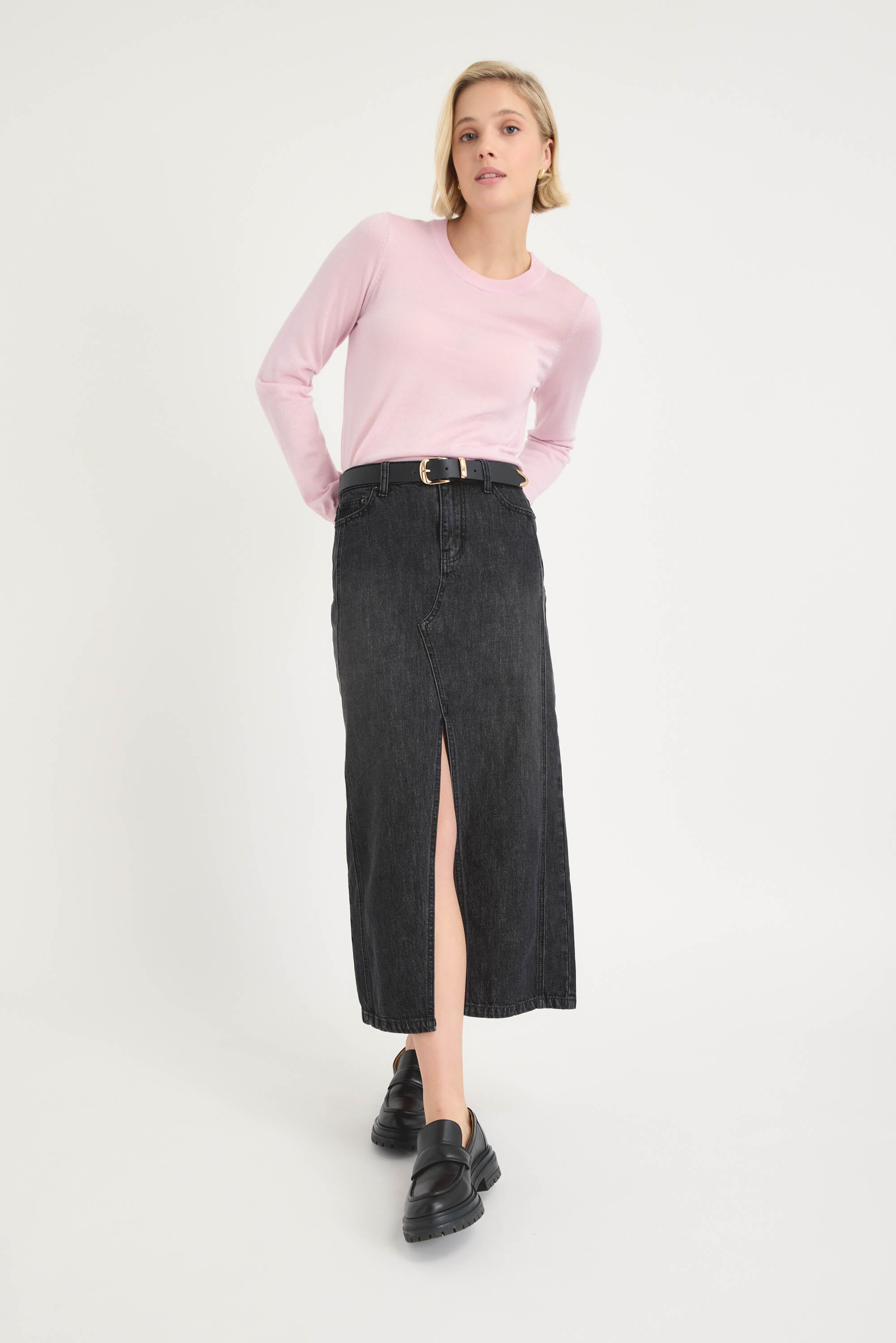 A woman poses against a plain white background wearing a light pink long-sleeve shirt and the Max Paris Maxi Denim Skirt in black, styled with a black belt and black platform shoes. Her hands are behind her back.