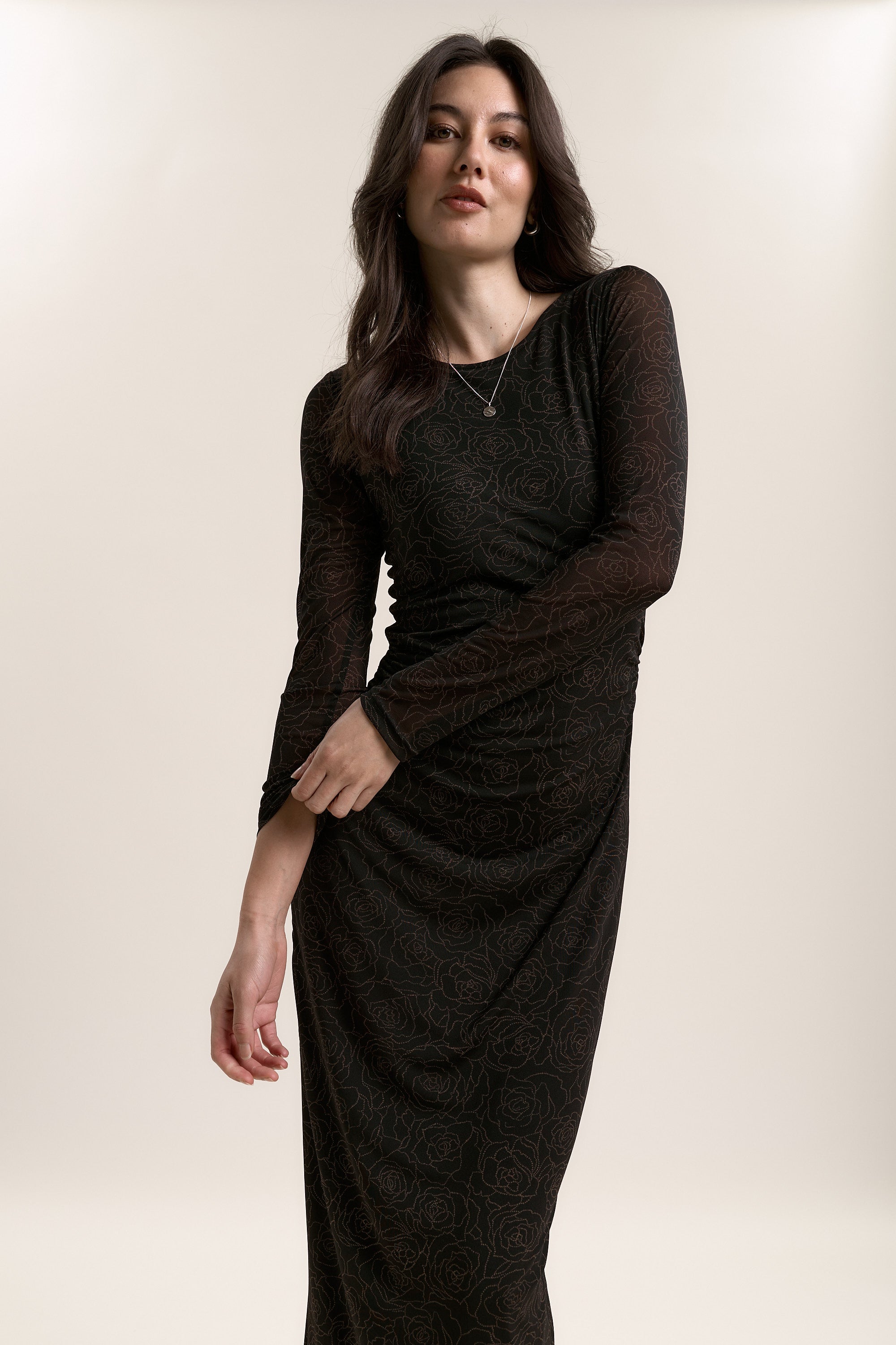 A woman with long dark hair models the Max Samara Long Sleeve Mesh Dress, featuring a subtle floral pattern, as she poses against a beige background with one arm bent across her waist and gazes slightly upward.