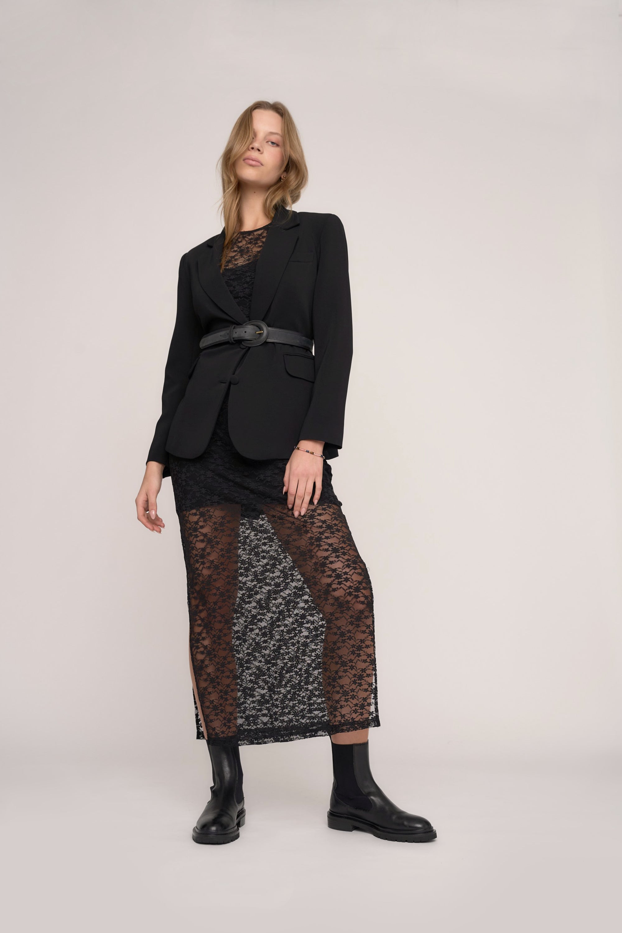 A woman with light wavy hair poses confidently in the Max Morgan Lace Dress—a sheer sleeveless lace dress with a visible slip, styled with a black belted blazer and black ankle boots against a plain background.