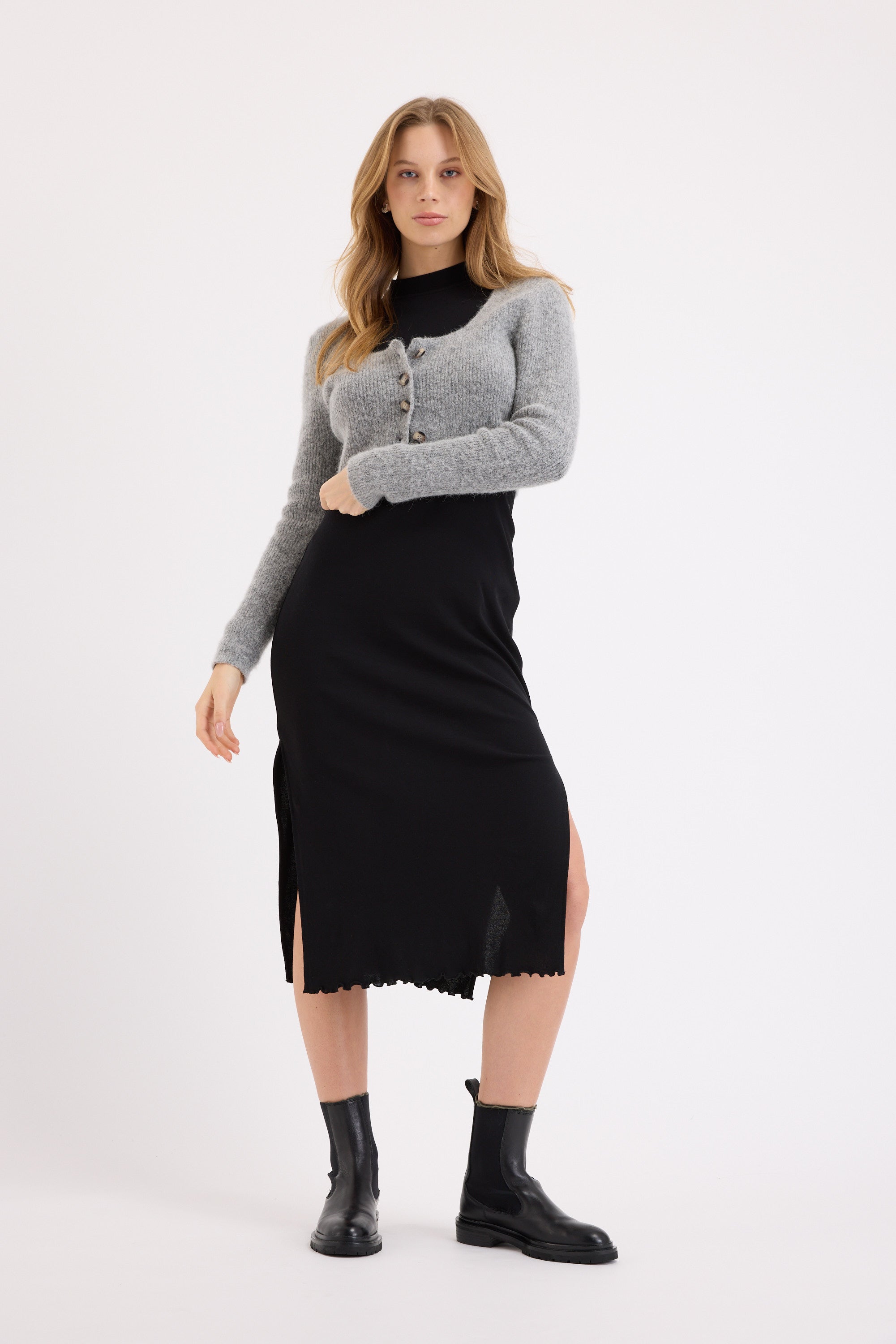 A woman poses confidently in the Max Nichole High Neck Layering Dress, styled with a cropped gray cardigan and black ankle boots against a white background. She stands with one hand on her hip, creating a chic winter look.