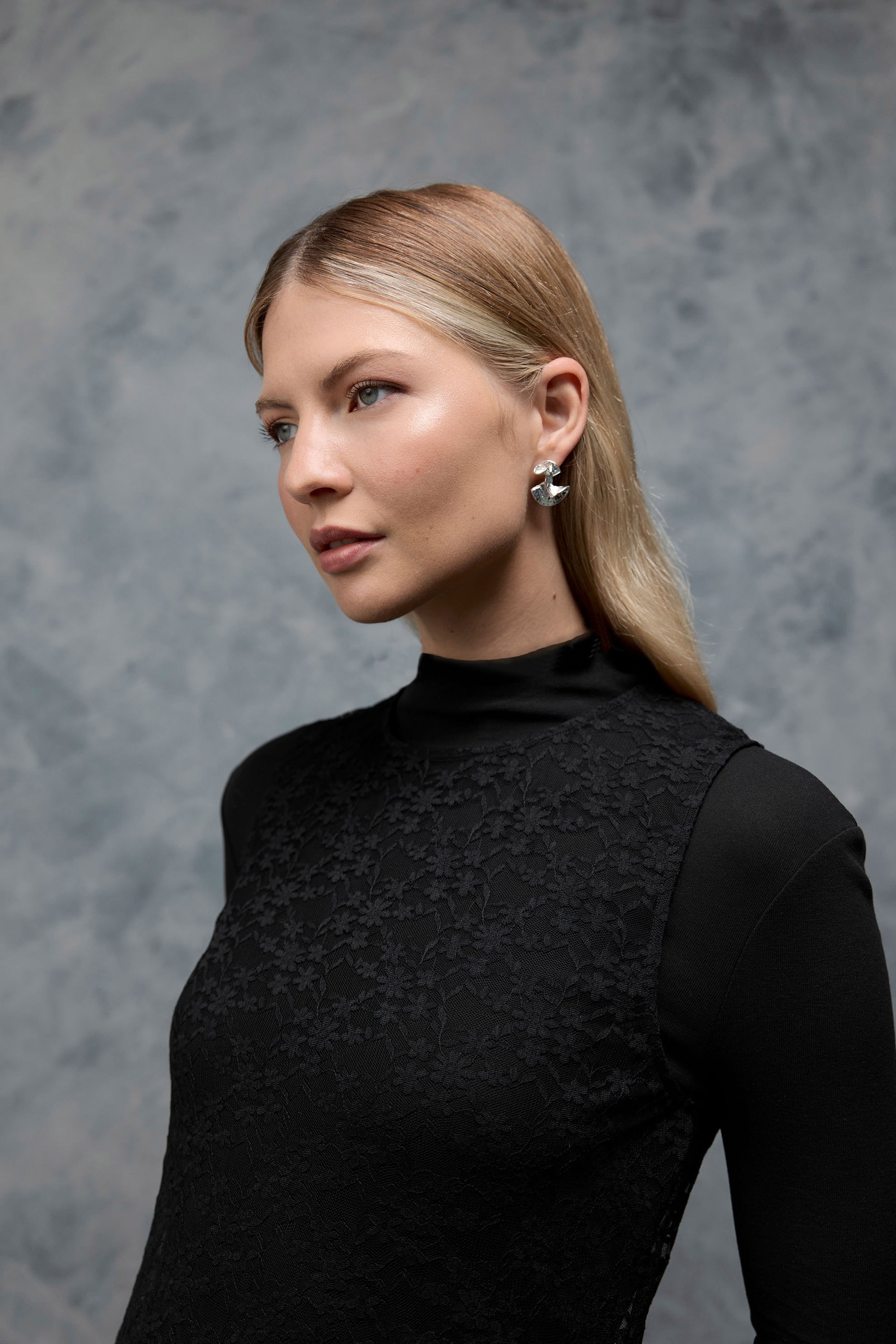 A woman with blonde hair wears the Max Nichole High Neck Layering Dress with lace sleeves and silver earrings, standing against a gray textured background and gazing thoughtfully to the side.