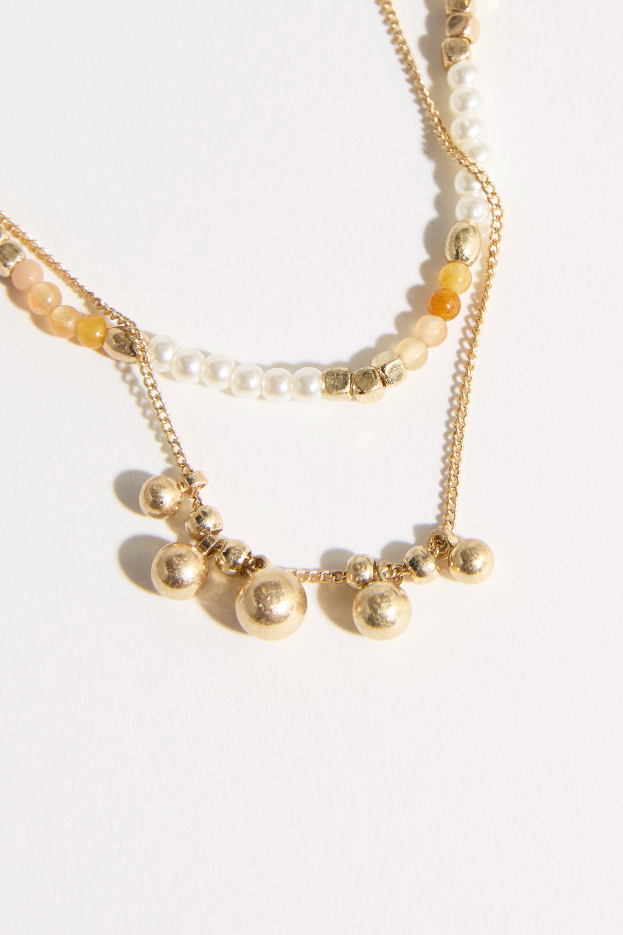 The Bela Sonder Kaia Necklace by Bela Sonder features small white, gold, and amber beads, paired with a gold pearl necklace with five round beads—elegantly displayed on a light background.