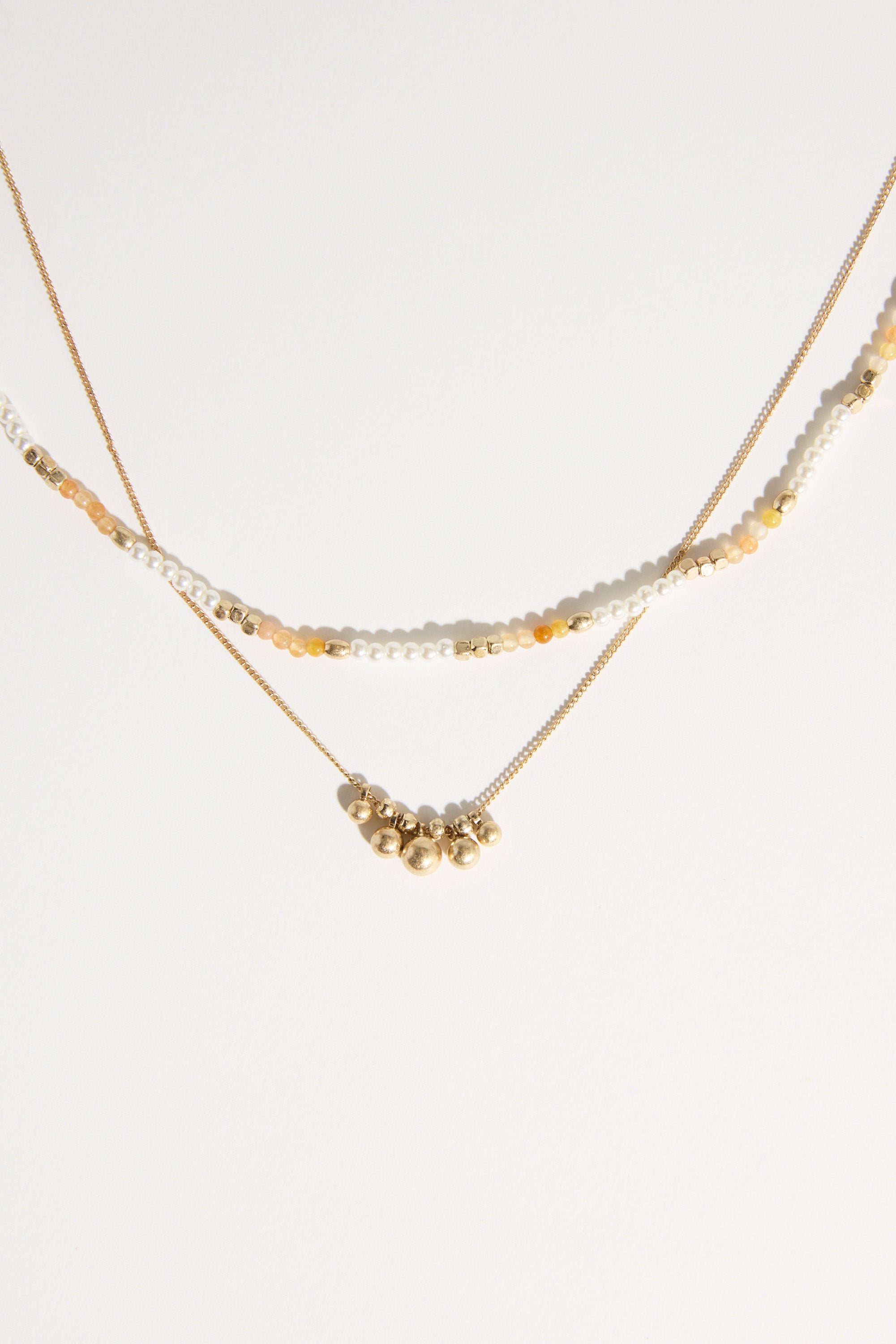The Bela Sonder Kaia Necklace features two layers: one with small white, gold, and yellow beads, and the other a gold chain with clusters of gold beads, shown on a white background. Brand: Bela Sonder.