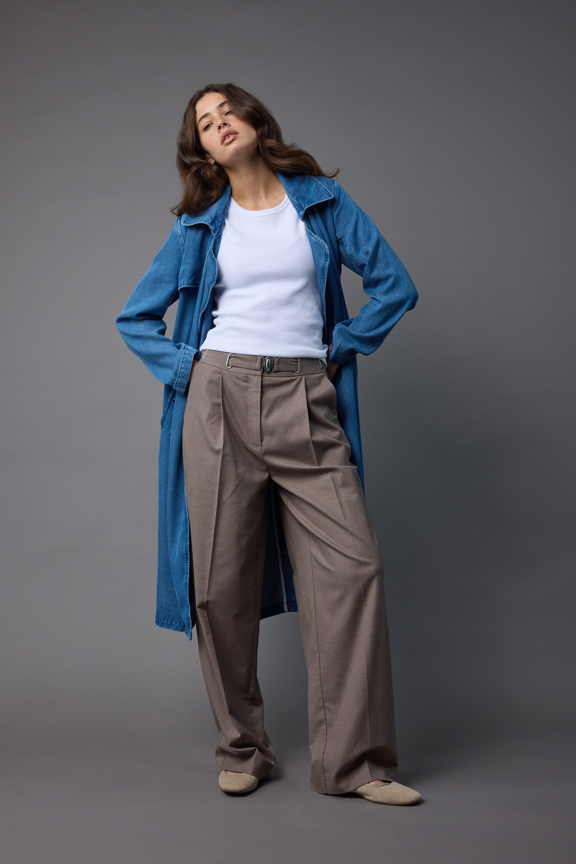 A woman poses against a gray background in a long blue denim coat, white ribbed shirt, Max's high-waist Sarah Buckle Detail Pant in brown, and beige shoes, with hands in her pockets and her head slightly tilted.