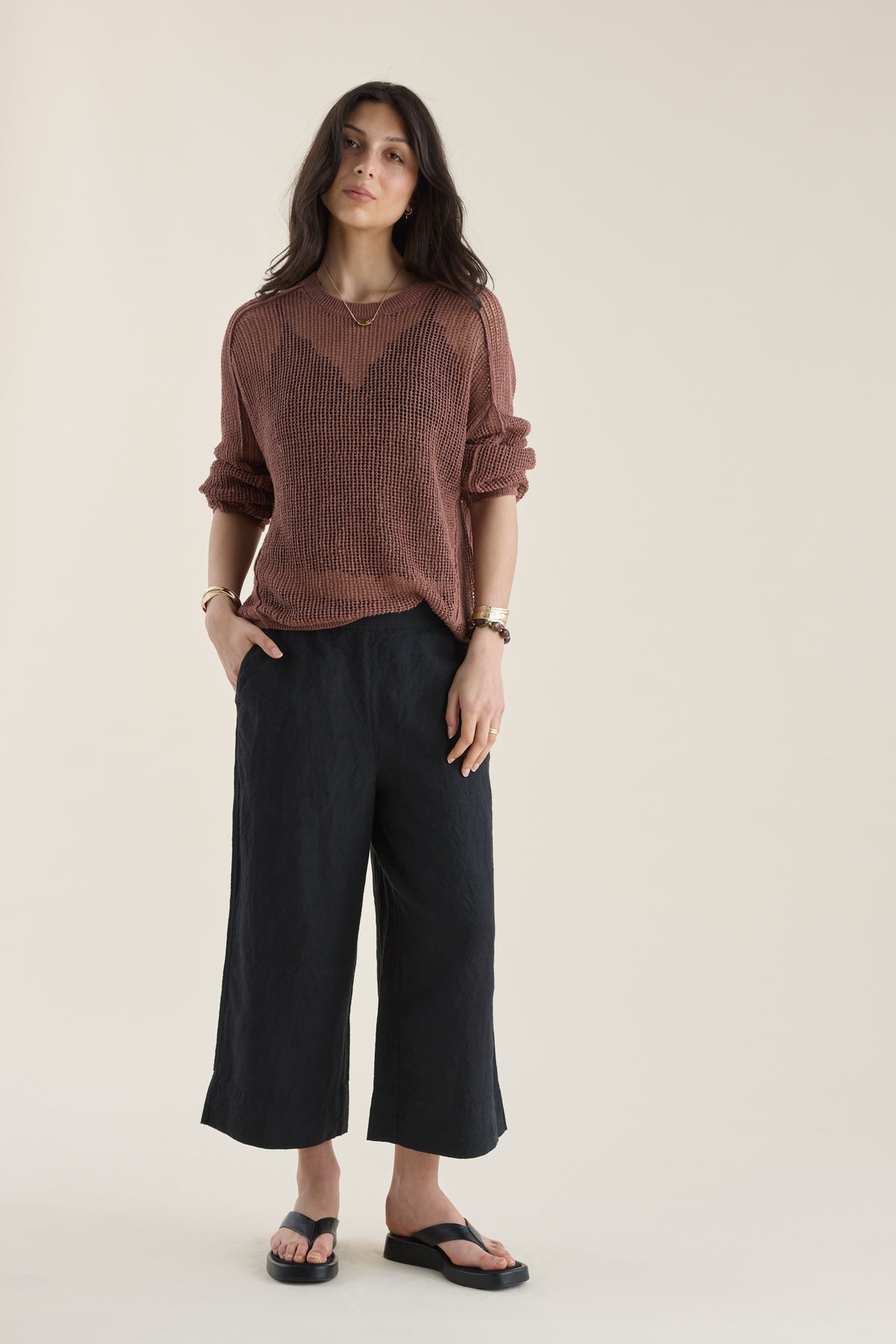 Woman in a brown sweater, Elementary High Tide Linen Crop Pants, and sandals against beige wall.