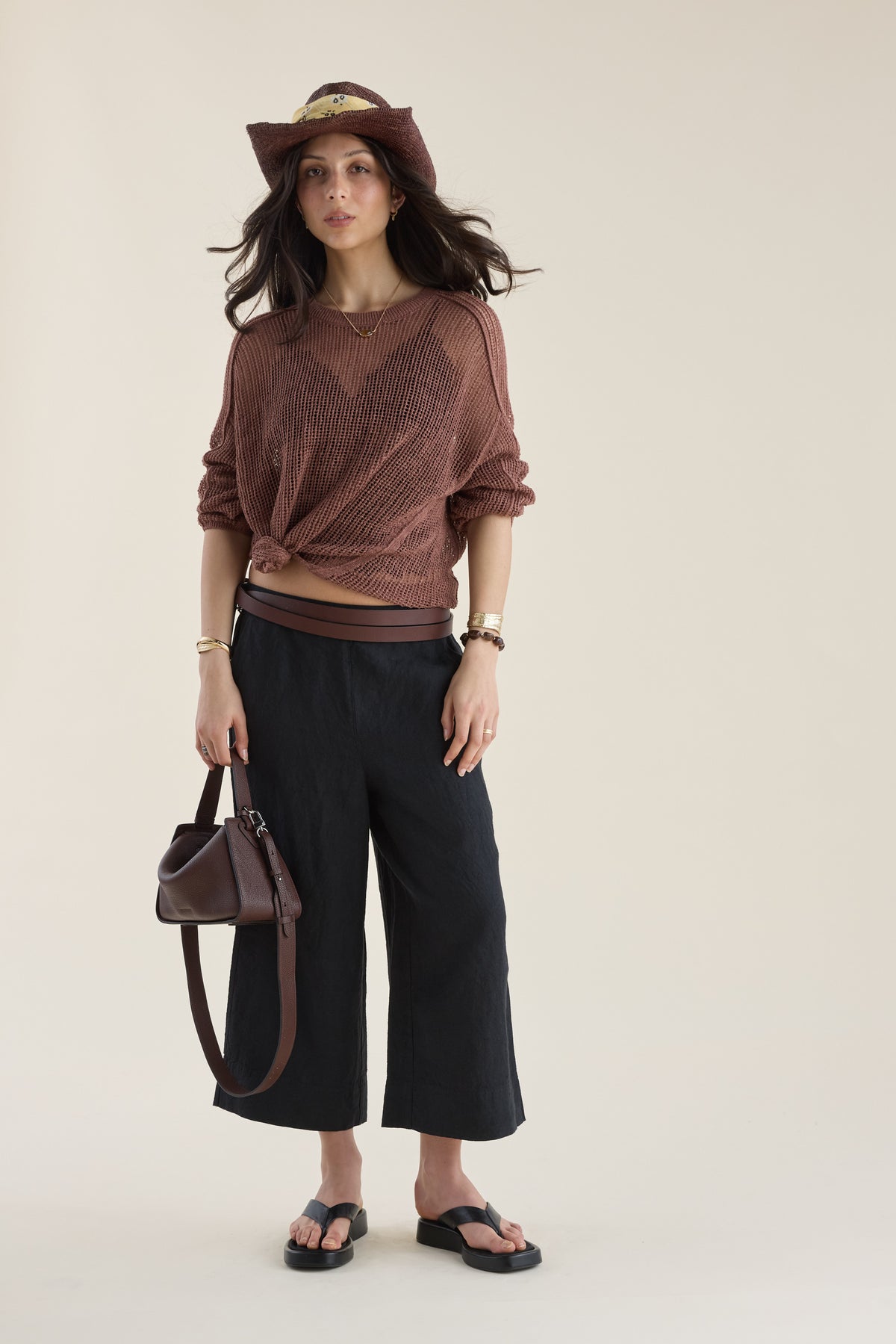 Woman in brown top, hat, Elementary High Tide Linen Crop Pants holds bag on neutral background.