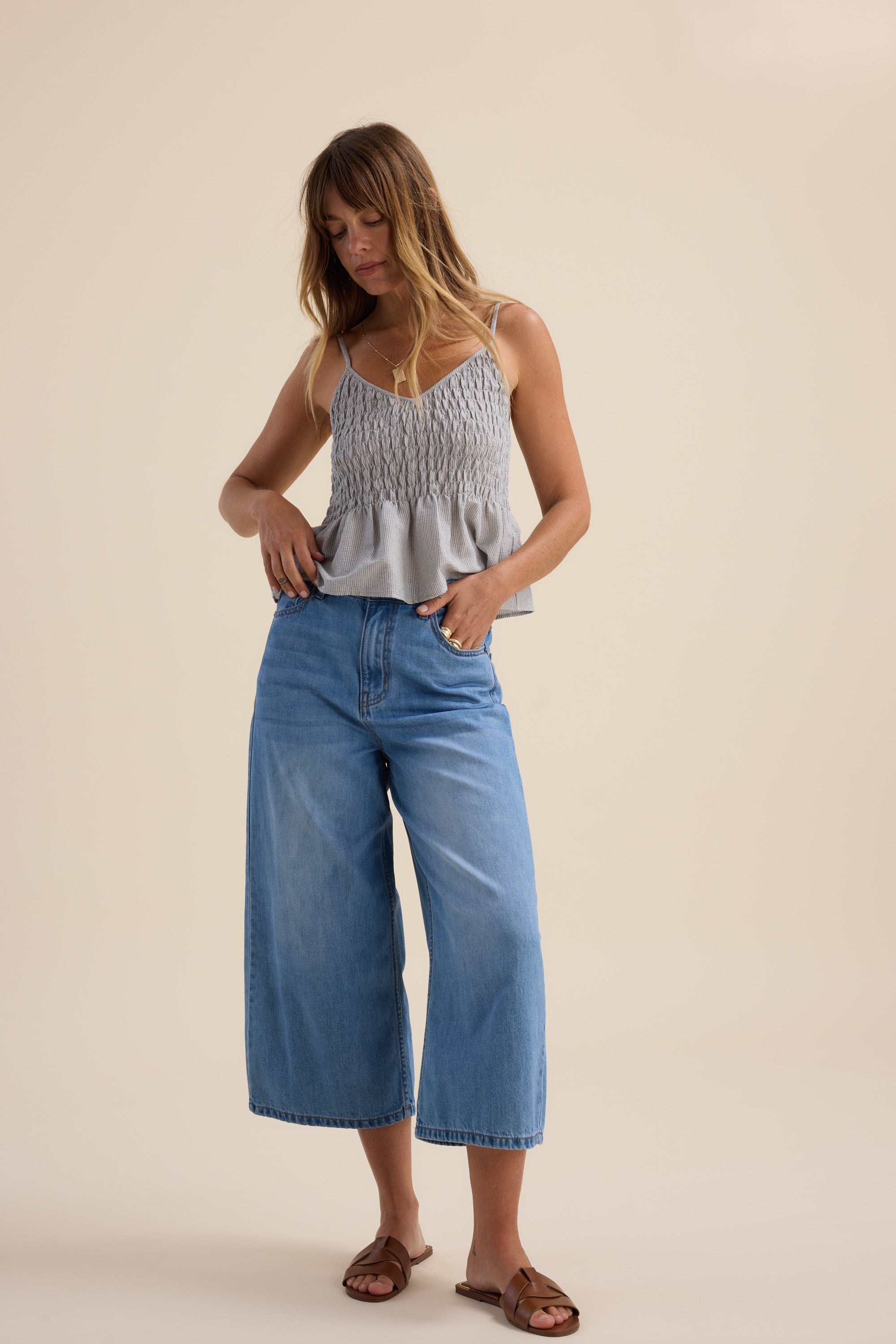 A woman with long, wavy hair stands against a beige backdrop, wearing Max's Derby Wide Leg Cropped Jeans in blue eco denim, a light gray sleeveless ruffle-hem top, and brown sandals. She glances down with one hand on her hip.