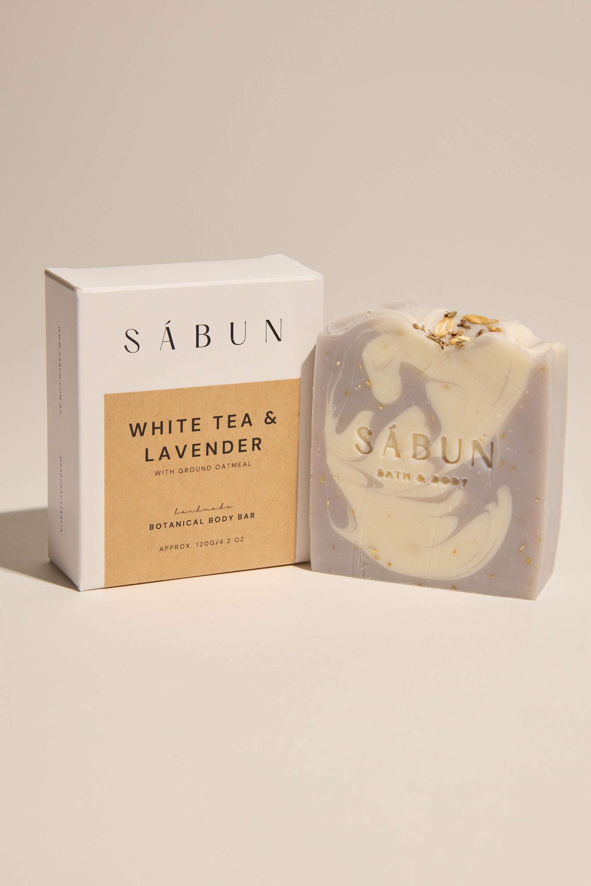 A Sabun Body Bar by Sabun, in White Tea & Lavender scent with ground oatmeal. The handmade Australian soap is light purple with a white swirl and specks, shown beside its box on a neutral background.