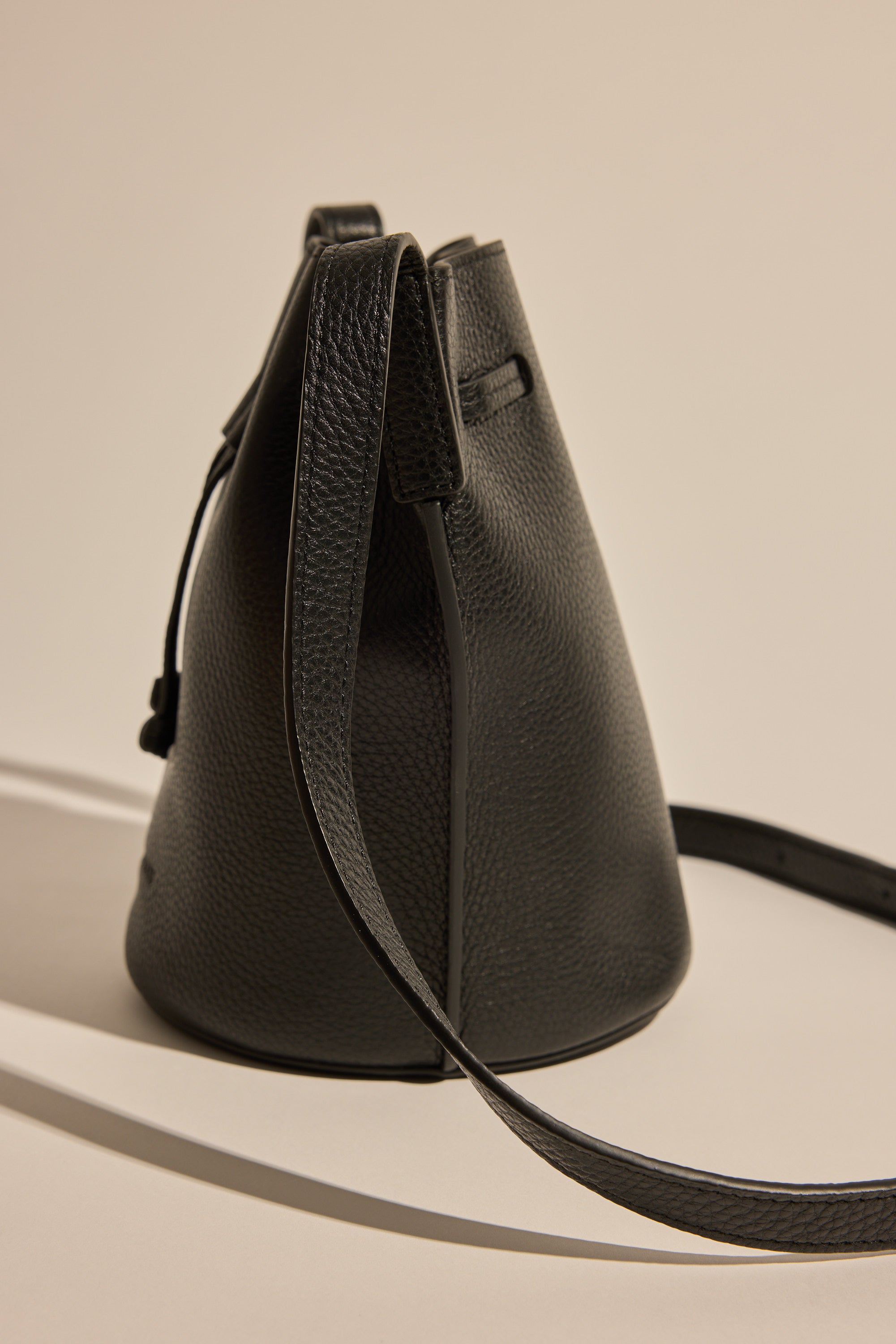 The Horse Bob Bucket by The Horse, a black grained leather bucket bag with a long crossbody strap, stands upright on a neutral background with soft shadows.