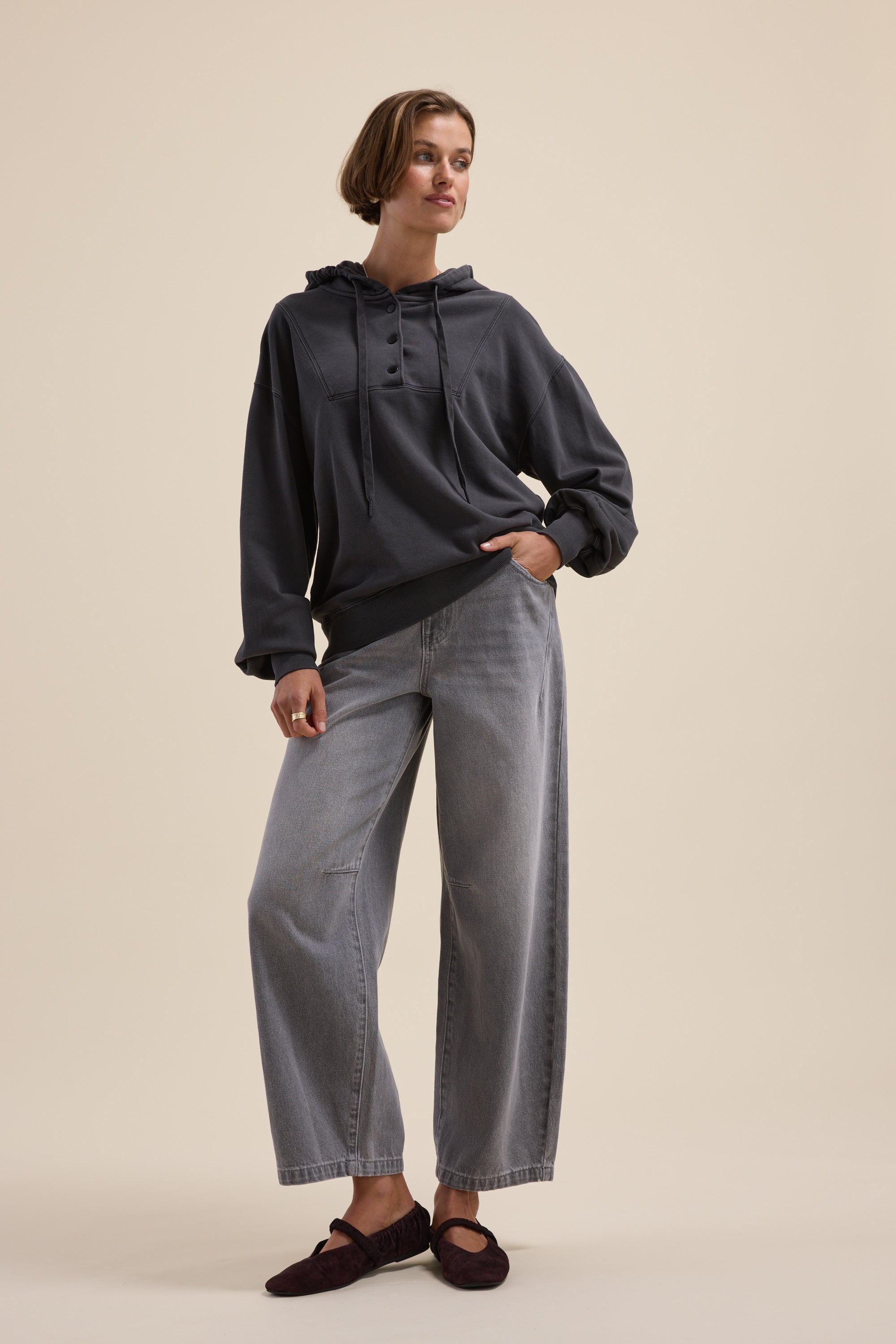 Woman in relaxed fit Elementary Henley Hoodie, loose gray jeans, and dark shoes on plain background.