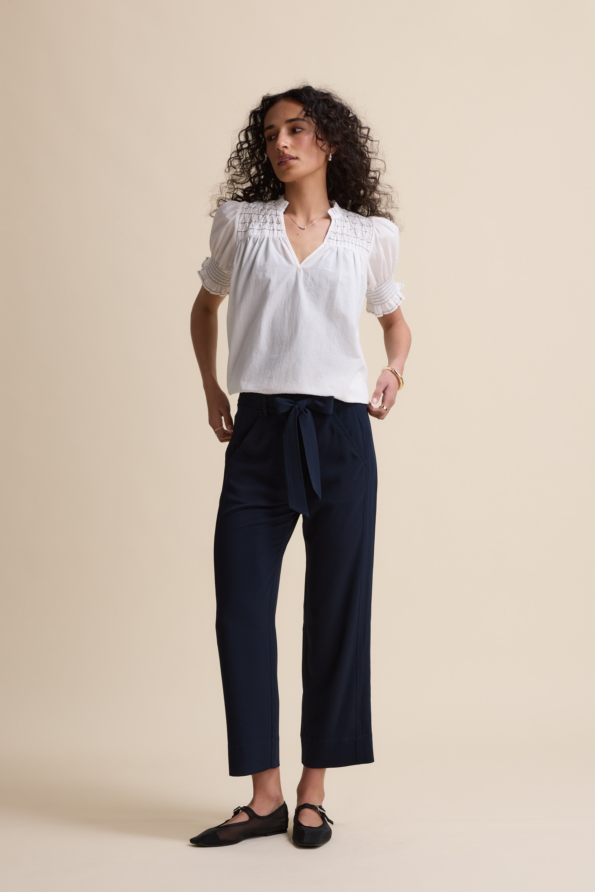 A woman with curly hair stands against a beige background, wearing Max's The Easy Care Belted Pant in dark blue. She pairs the high-rise, wide-leg pants tied at the waist with a white textured blouse and black shoes, looking to the side.