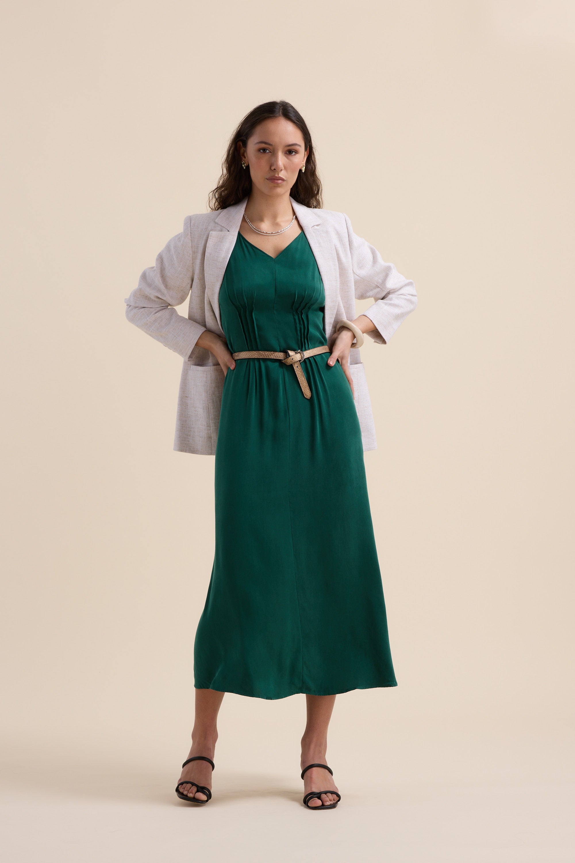 A woman stands against a beige background, confidently wearing the Max Cara Pleat Detail Dress—green with a cinched waist—styled under a white blazer, black heeled sandals, and a necklace. She has wavy brown hair and her hands on her hips.