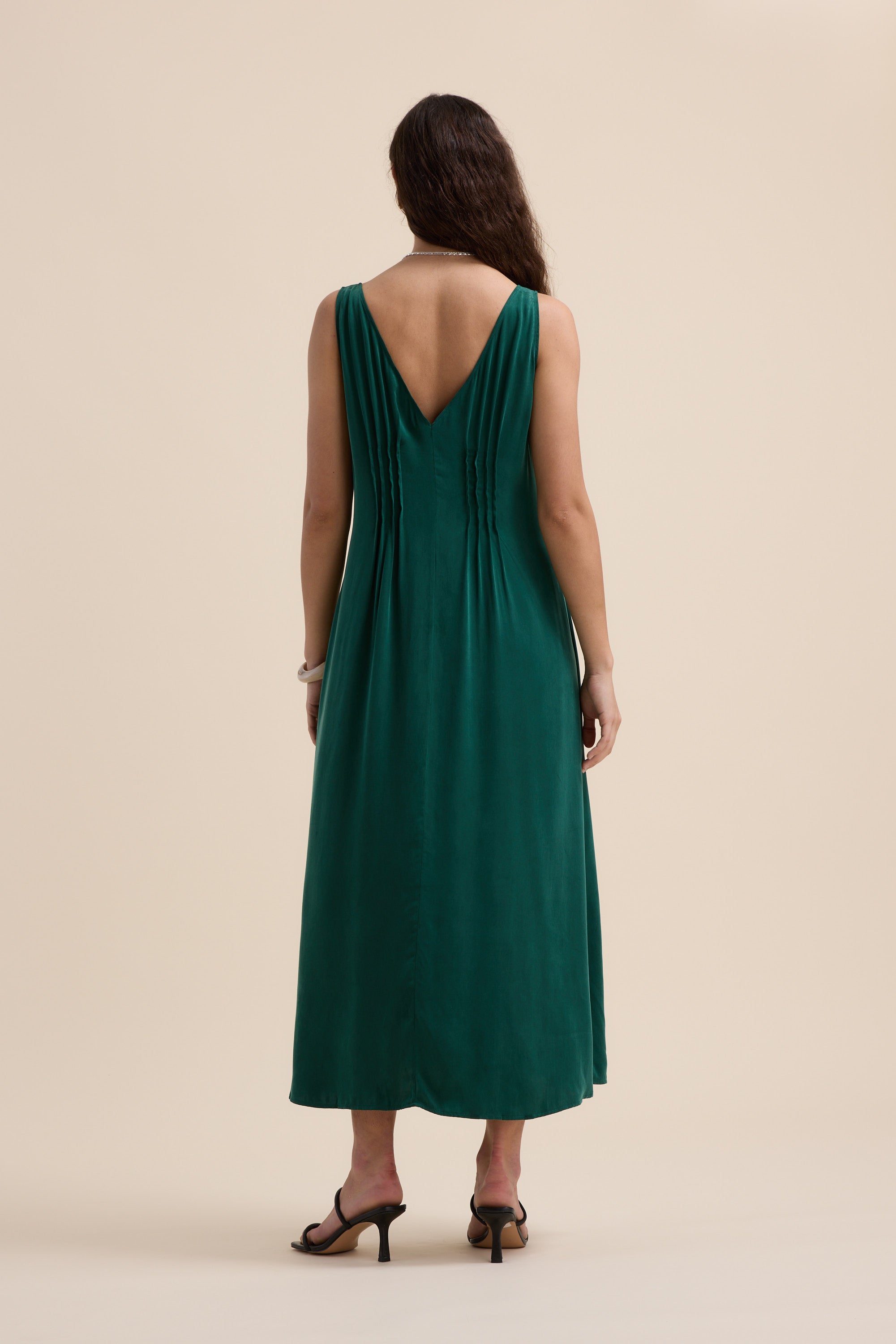 A woman with long brown hair, seen from behind, wears the Max Cara Pleat Detail Dress—a sleeveless green maxi dress with a deep V-back, cinched waist, and pleats. She stands on a neutral background in black heeled sandals.