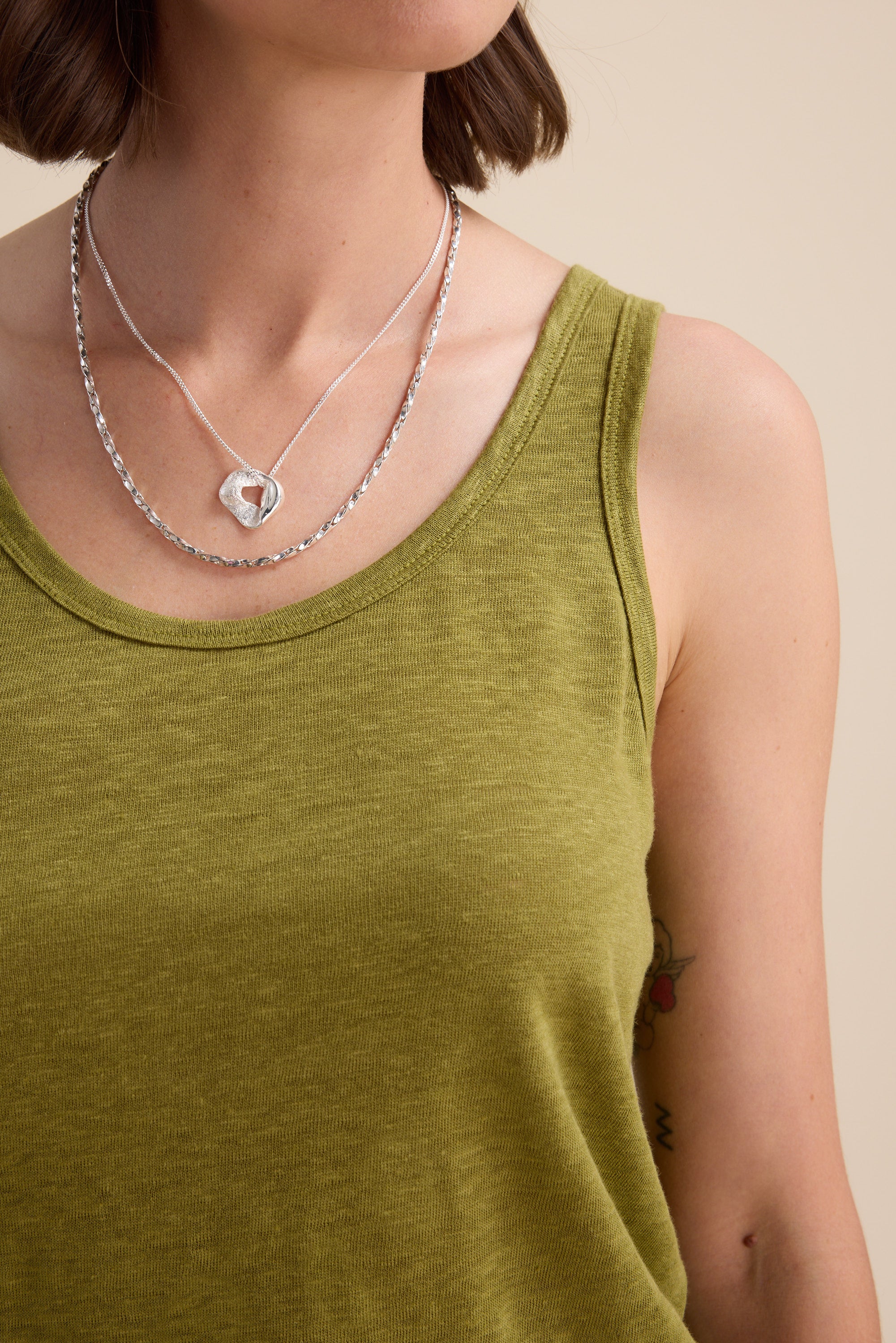 A woman wears the Elementary Linen Jersey Tank by Elementary in light green, paired with two silver necklaces—one featuring a geometric pendant. A small floral tattoo is visible on her upper arm, set against a plain, neutral background.