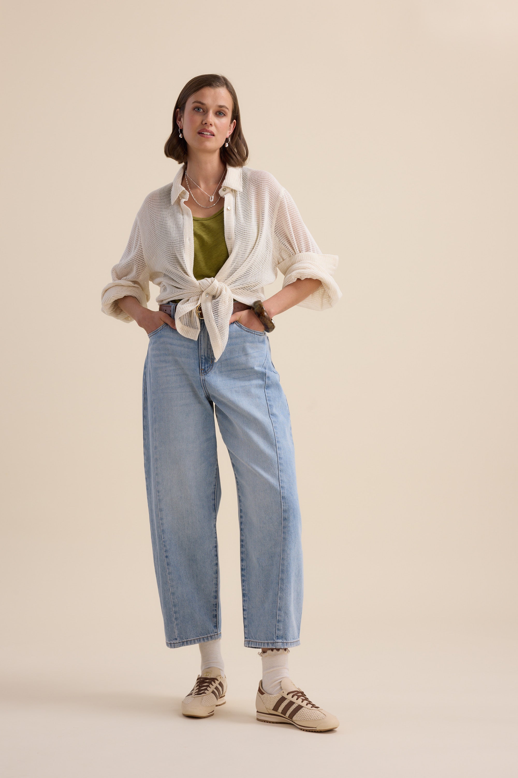 A woman with short hair stands against a beige background, wearing the Elementary Linen Jersey Tank by Elementary in green under a sheer white shirt, paired with high-waisted light blue jeans, beige sneakers, white socks, earrings, and a watch.