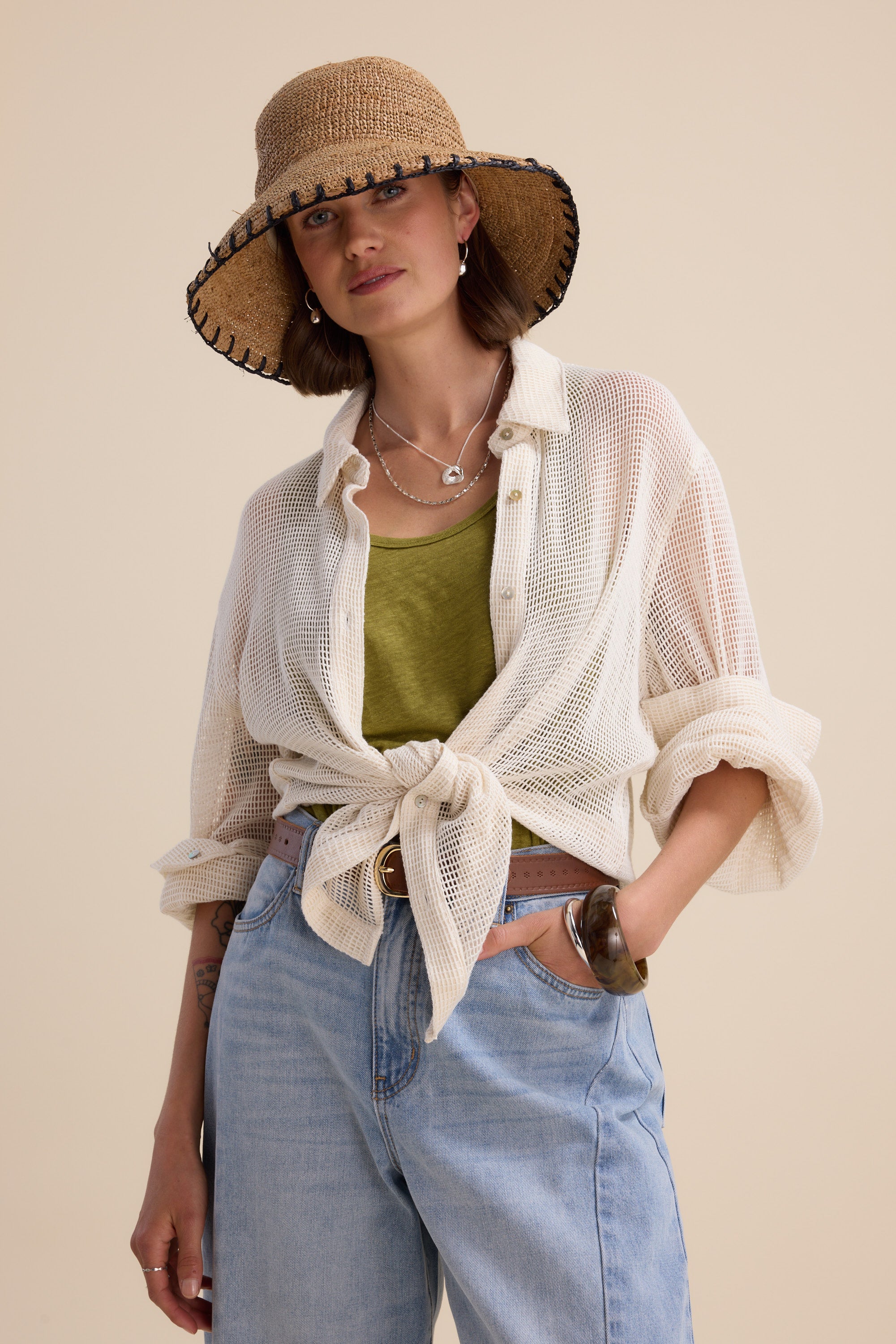 A woman stands against a beige background wearing the Elementary Linen Jersey Tank by Elementary, paired with a straw hat, sheer white button-down shirt tied at the waist, light blue jeans, and layered necklaces.