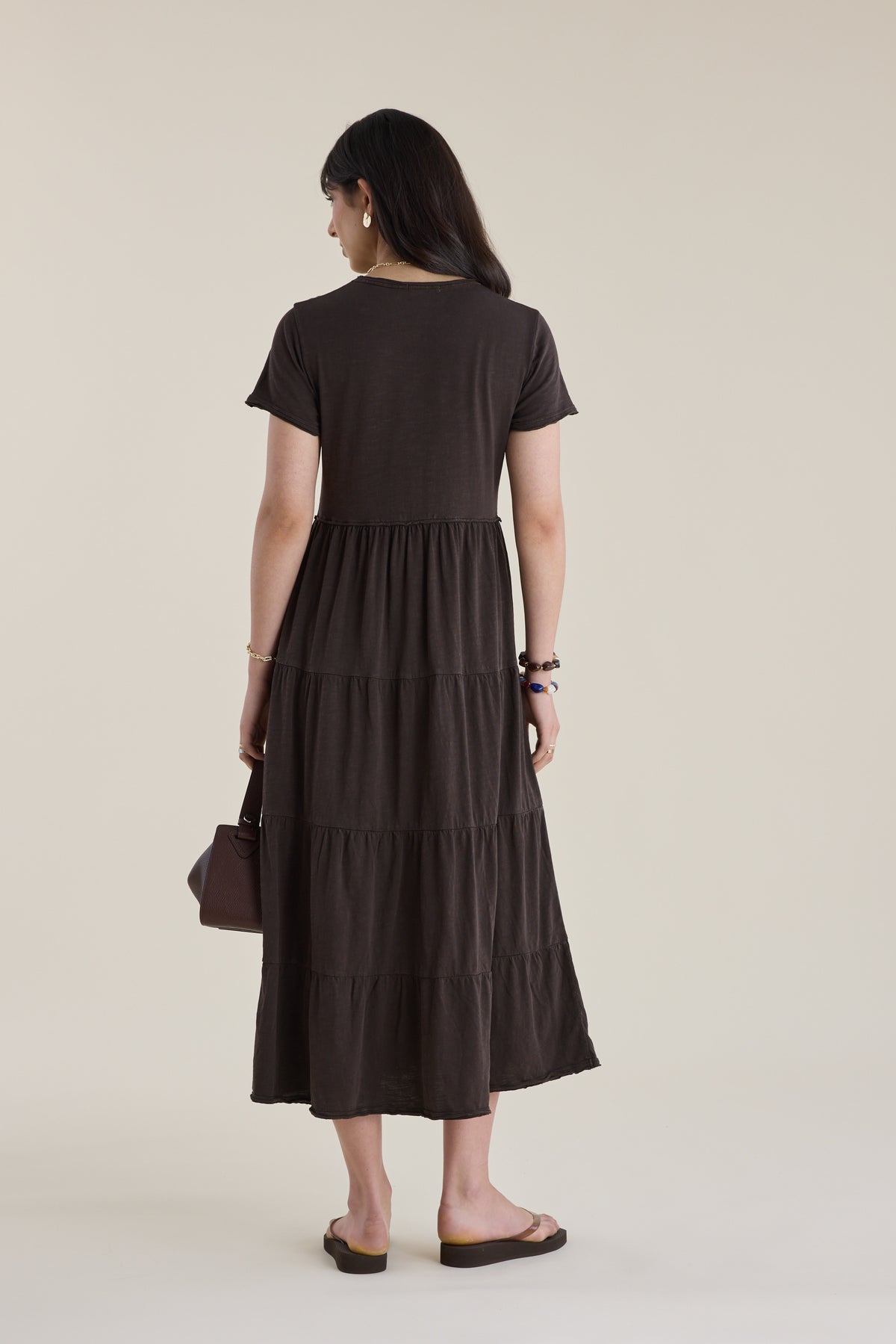 Woman in Elementary Jersey Tiered Dress, holding bag, stands with her back to the camera.