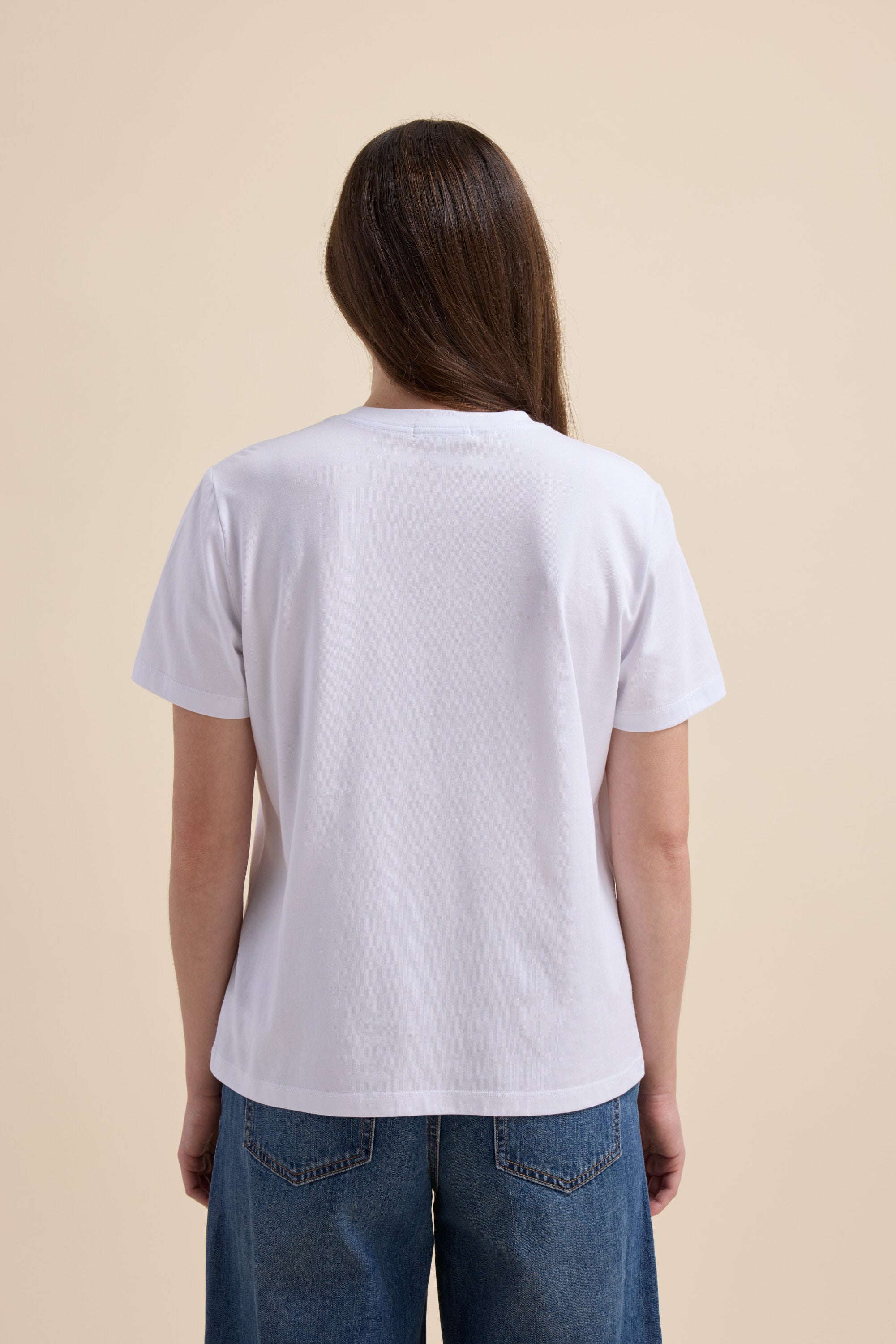 A person with long brown hair in a Max Good Earth Cotton Crew Tee and blue jeans, seen from behind.