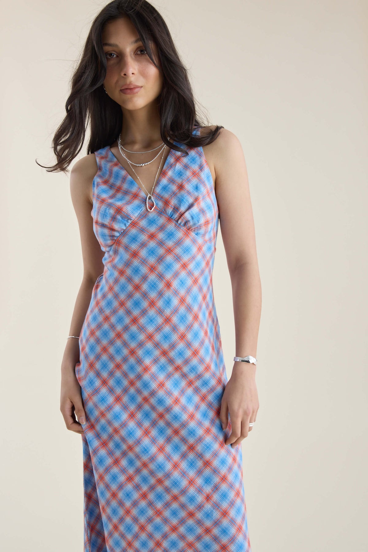 Woman in Max Nina Bias Cut Dress, blue/red checks, layered necklaces, plain backdrop.