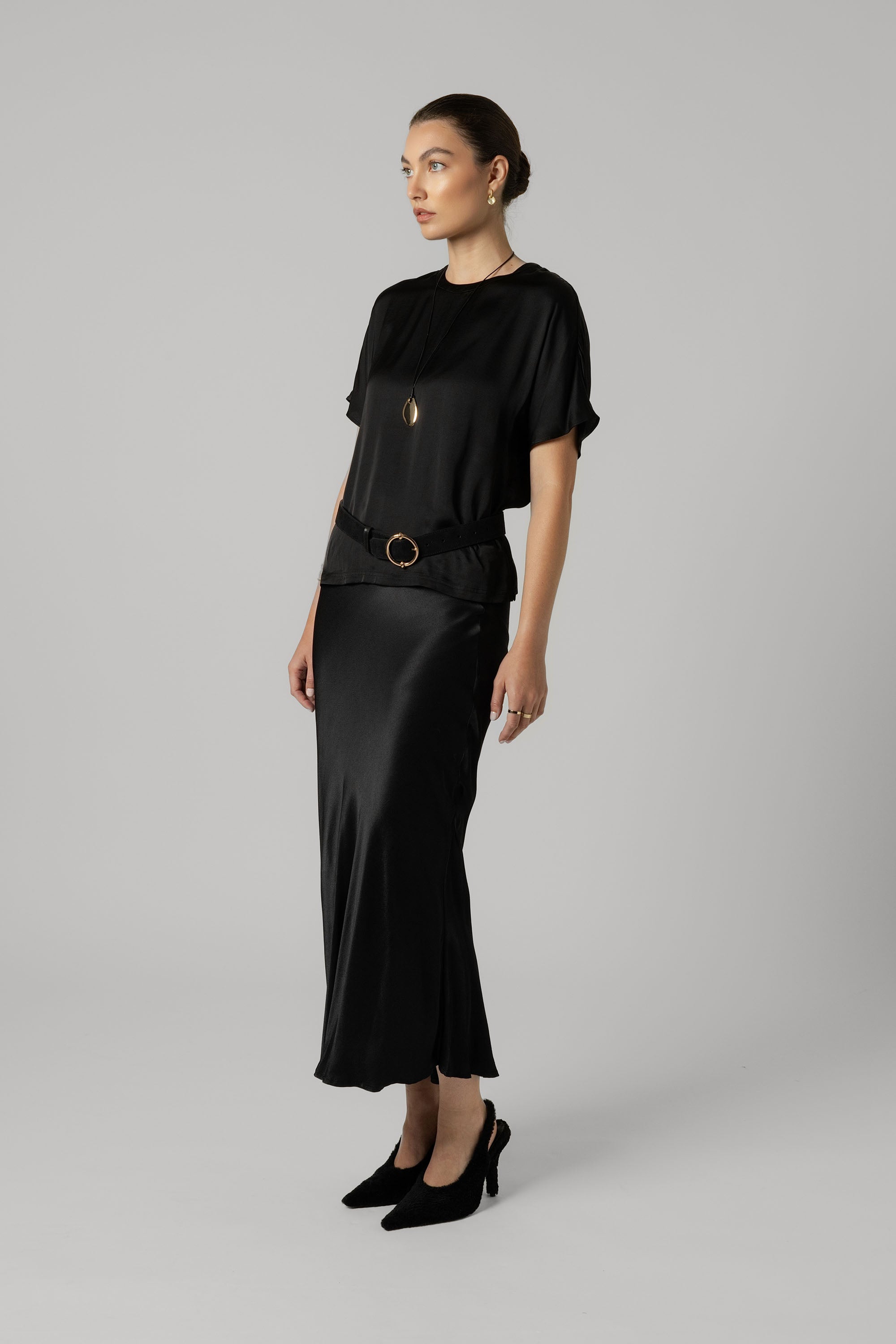 Woman wears Max’s The Perfect Slip Skirt in black with a matching top and heels, against gray.