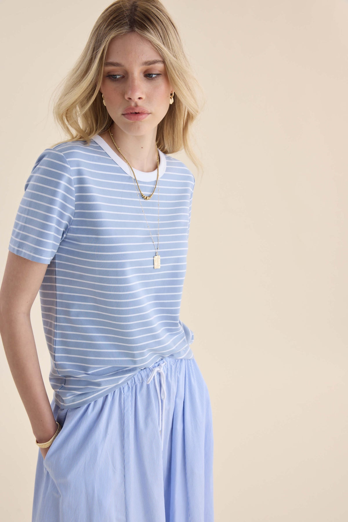 Woman in Max’s Perfect SS Crew Neck Tee, relaxed fit skirt, looking down, hands in pockets.