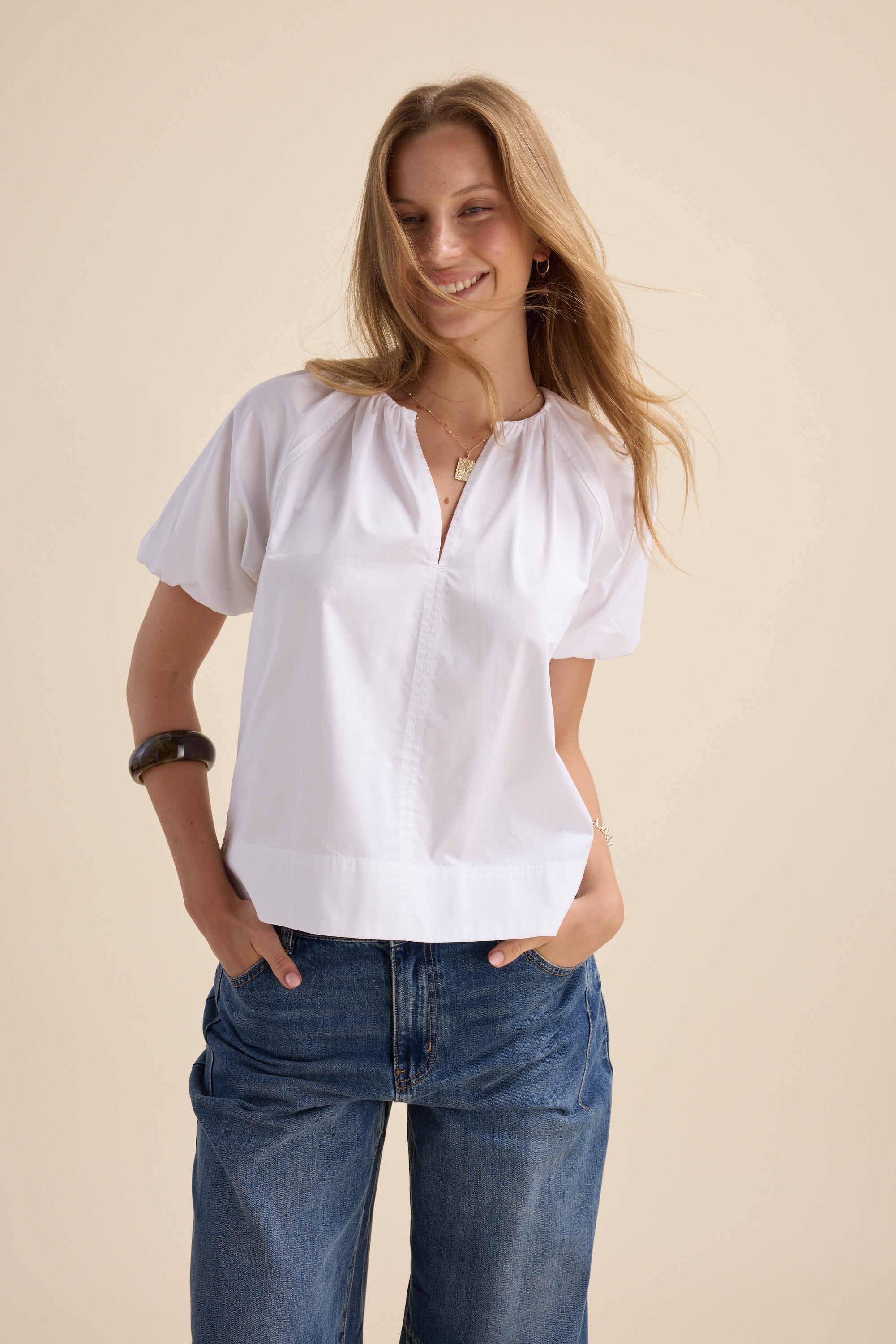 Woman smiles in Max’s The Poplin Blouse with puff sleeves, hands in pockets, beige background.