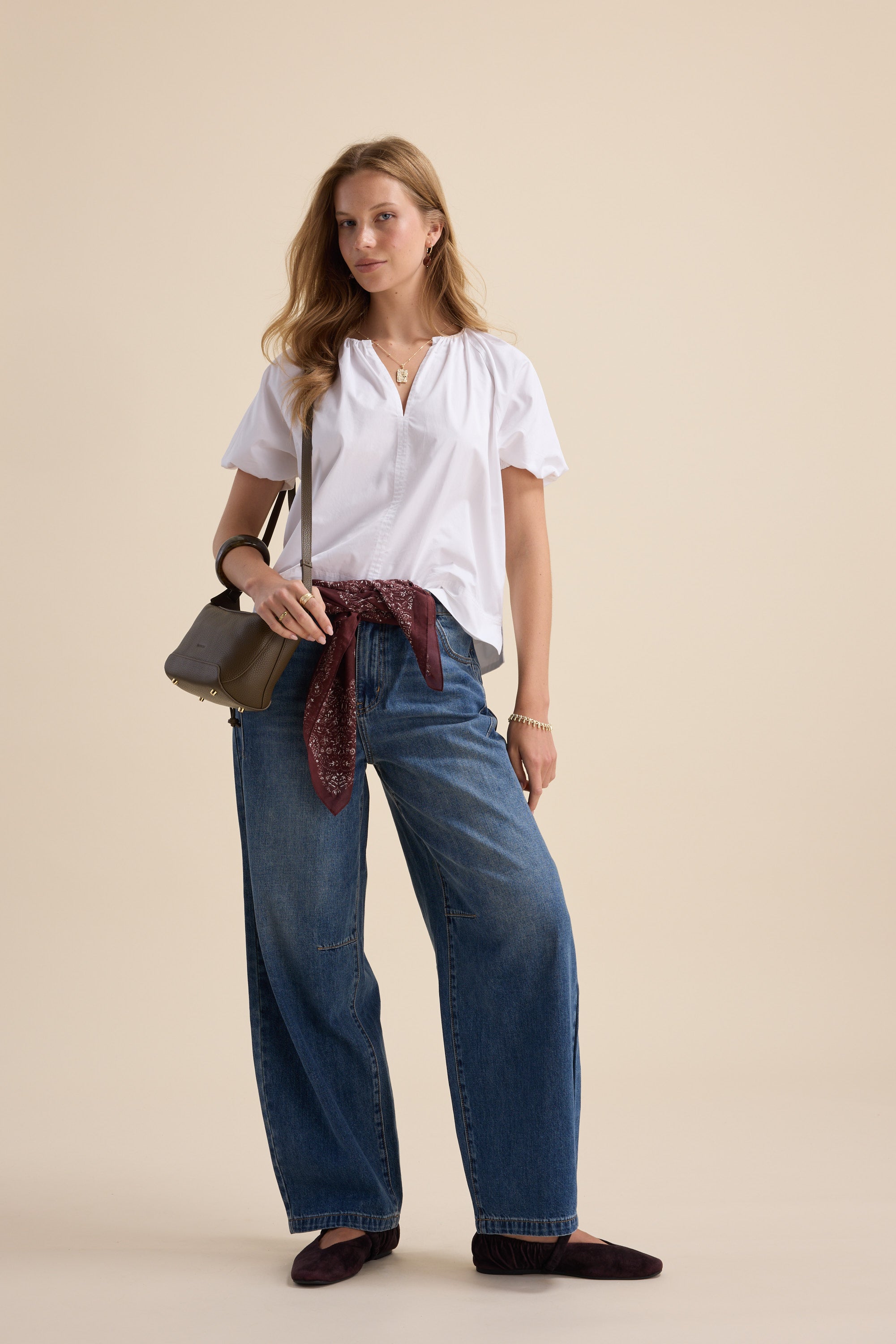 Woman wears Max’s The Poplin Blouse, wide jeans, bag, and maroon scarf on a beige background.