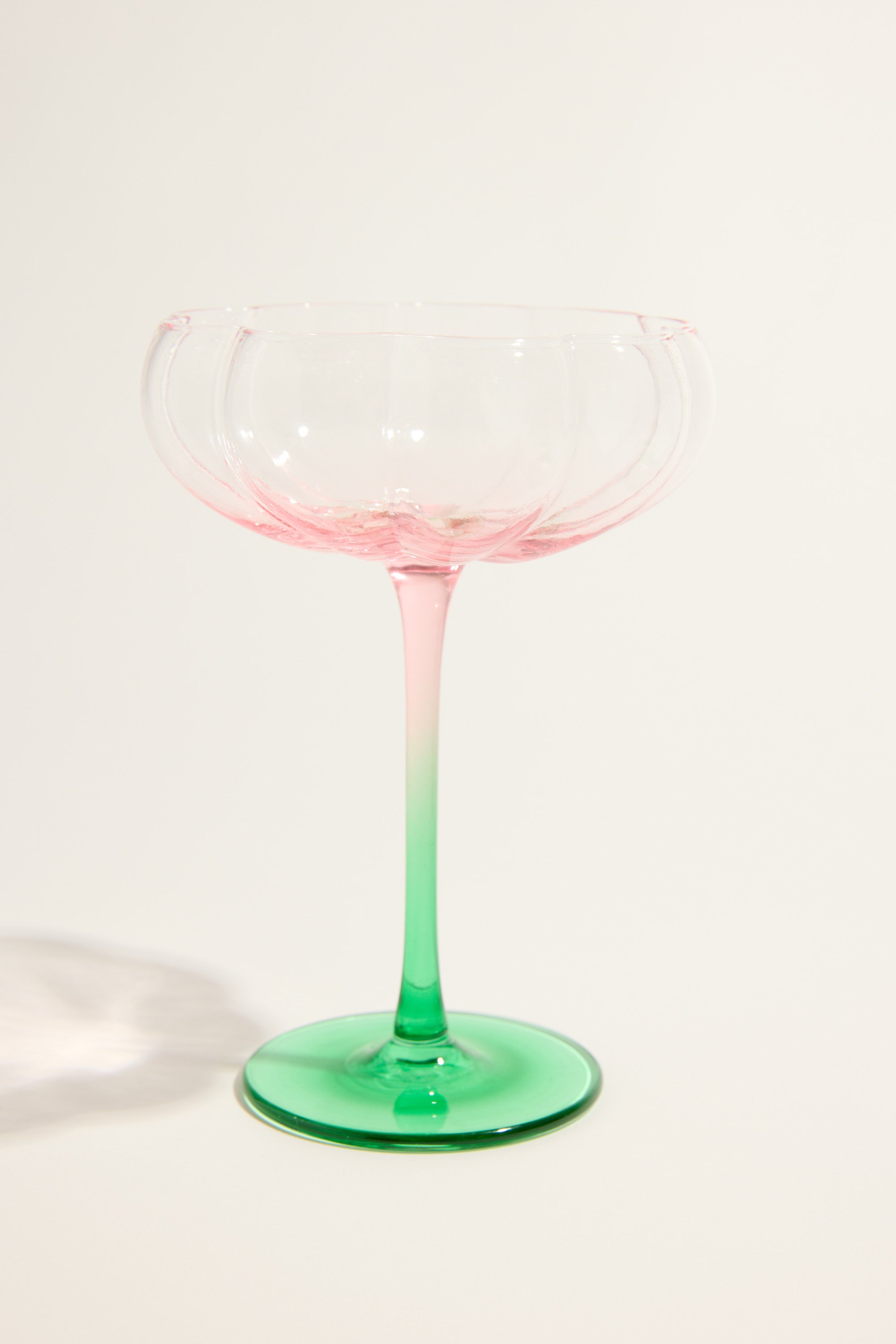 A delicate, empty coupe from the Homelab Petal Cocktail Glass Set by Homelab, featuring a pink bowl, green stem, and unique petal design against a light background. The elegant gradient enhances the charm of this glassware set.
