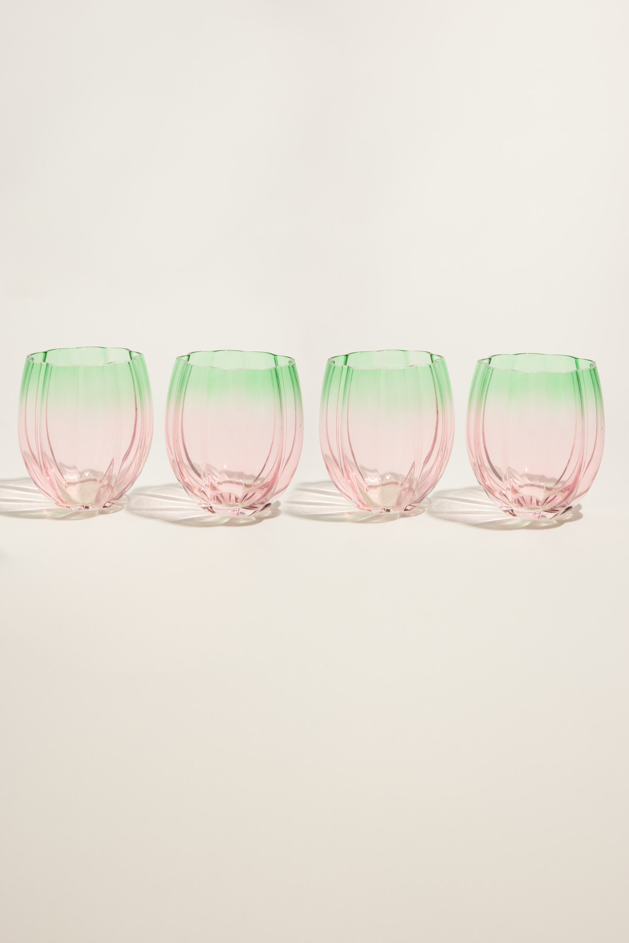 The Homelab Petal Stemless Glass Set by Homelab features four rounded glasses with pink bases and green rims, neatly arranged on a light background. The unique glassware casts gentle shadows across the surface.