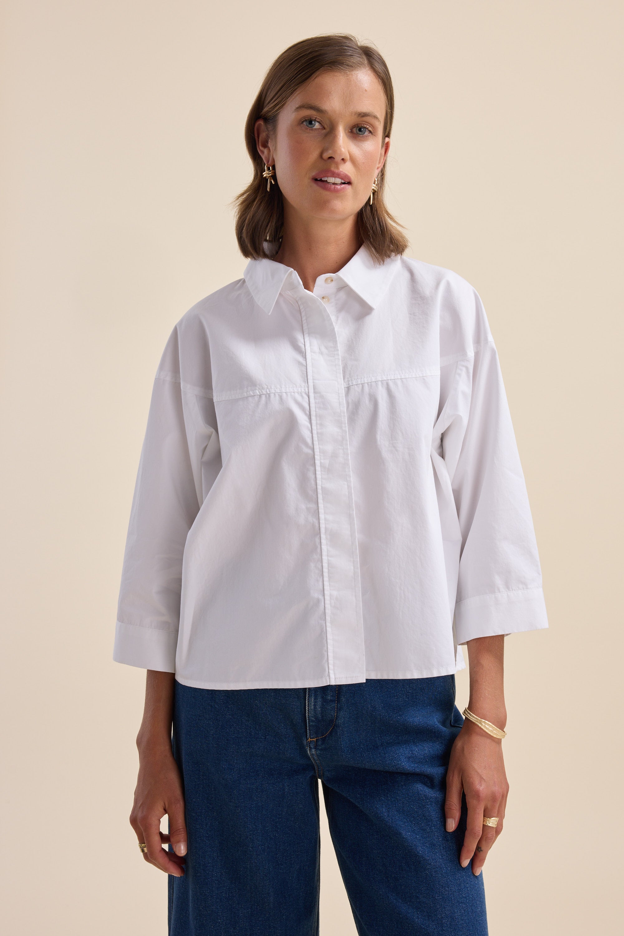 Woman in an Elementary Yoke Detail Shirt and blue jeans stands against a beige background.