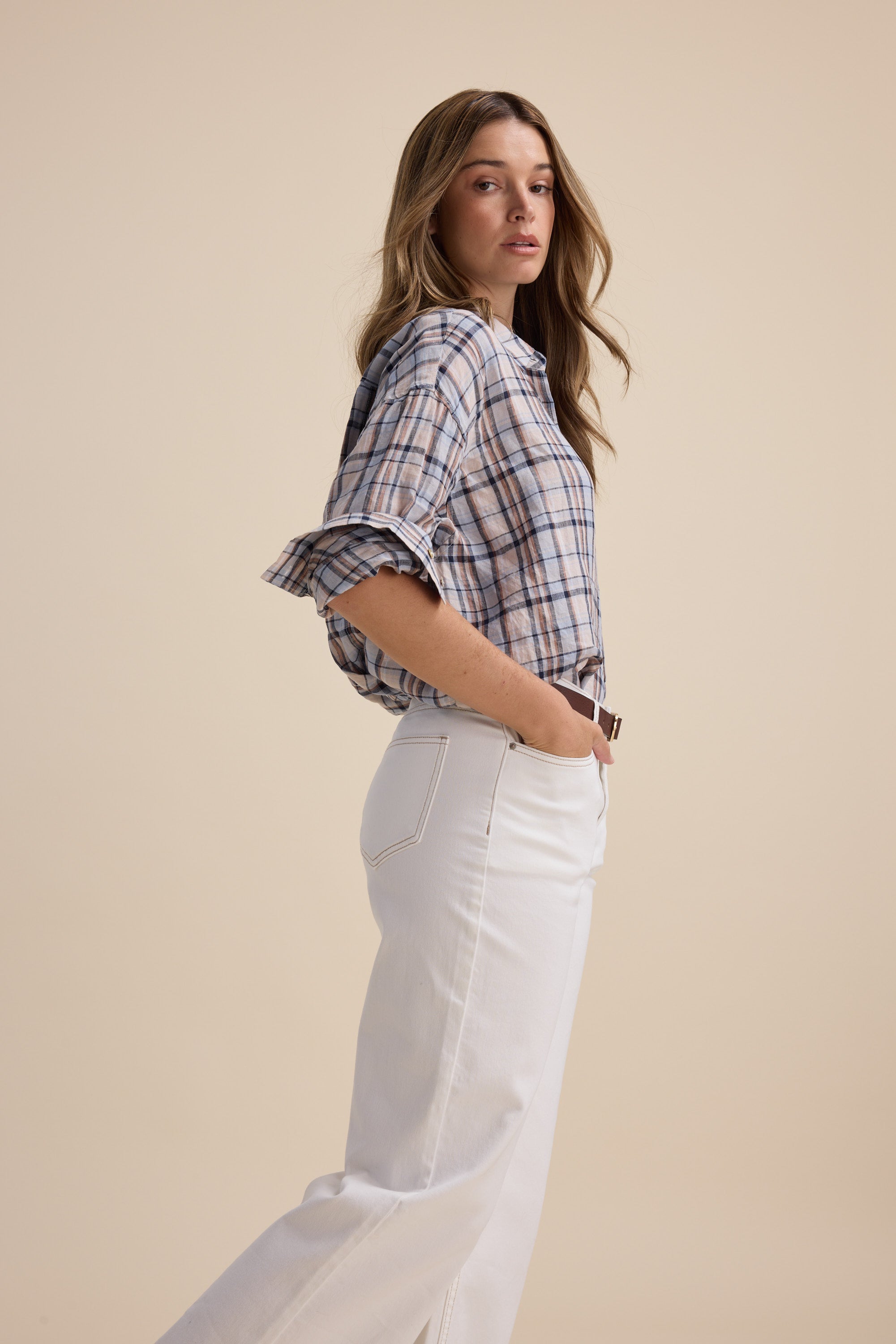 Woman wears Elementary Oversized Shirt and white pants, standing sideways with hands in pockets.