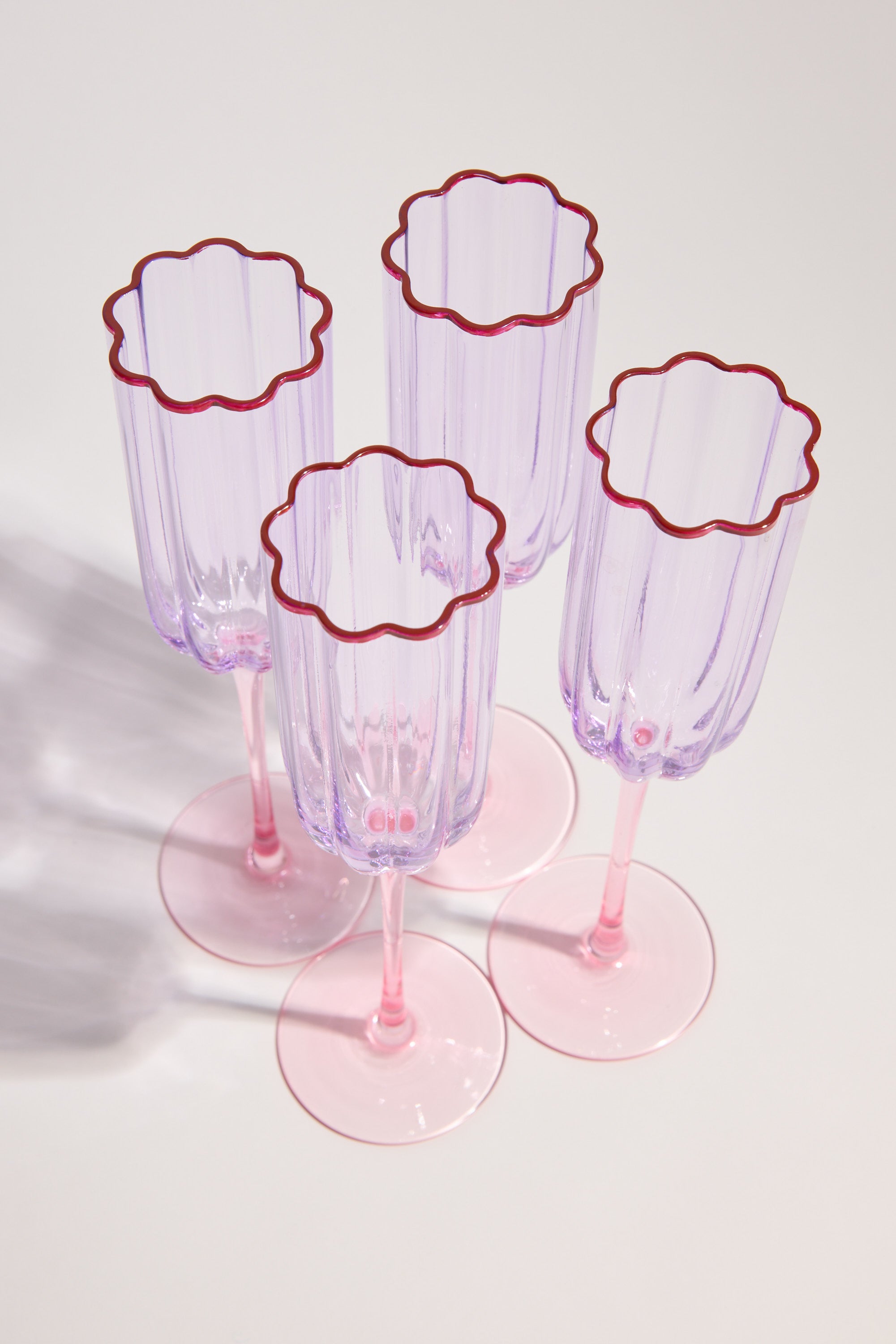 The Homelab Petal Champagne Flute Set by Homelab features four translucent pink glasses with scalloped, red-outlined rims, elegantly arranged on a light surface—an enchanting glassware set ideal for any celebration.